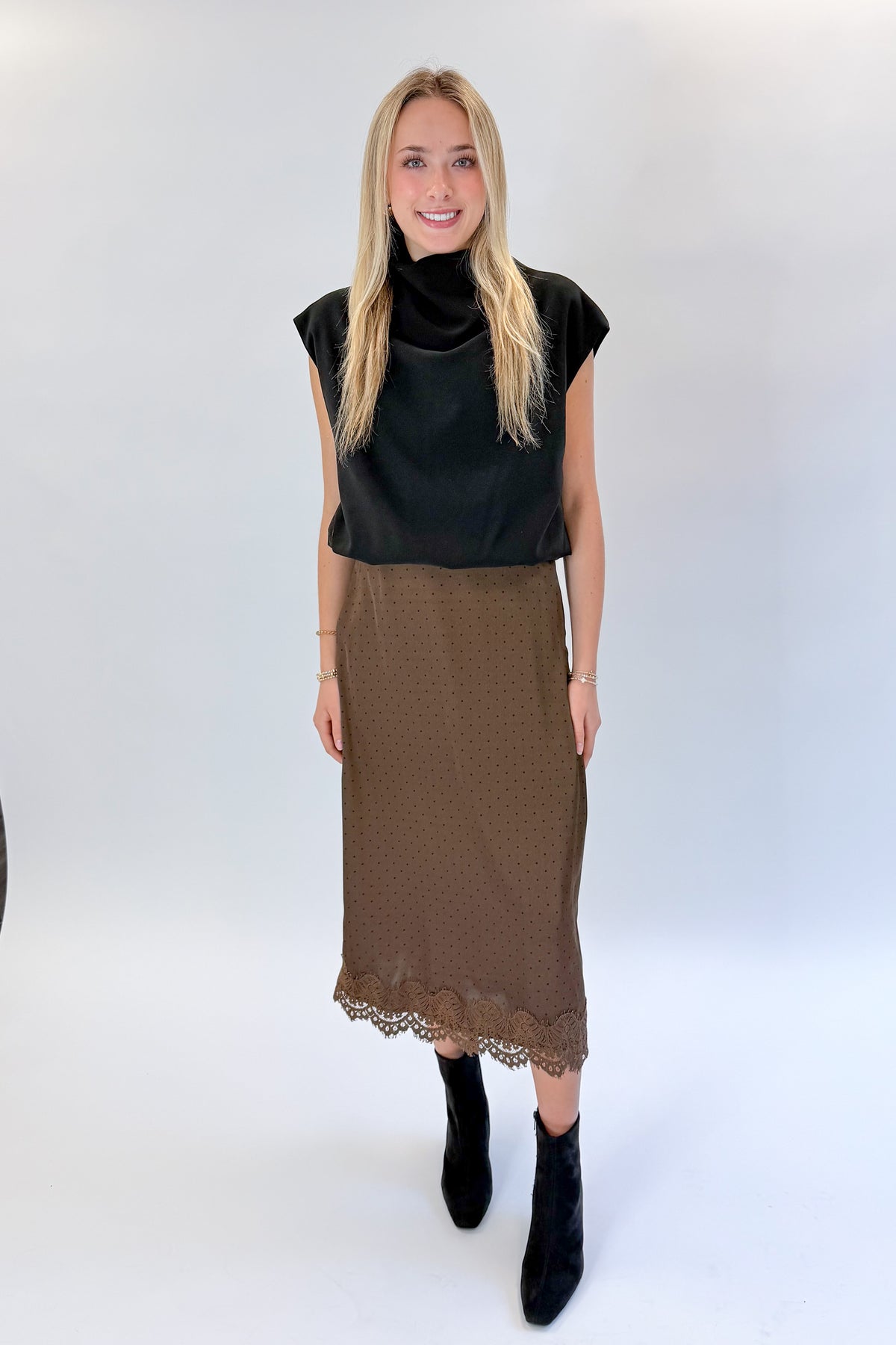 Front view of woman wearing Swagger Boutique’s Brown Polka Dot Lace Lined Satin Skirt by Entro with Black Satin Cowlneck Sleeveless Top against white studio background.