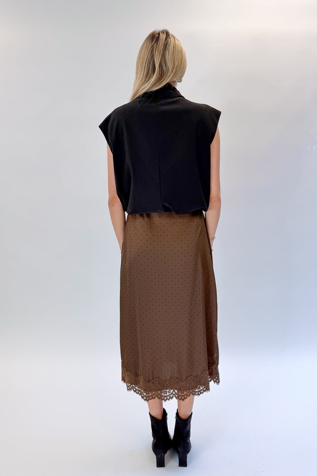 Back view of woman wearing Swagger Boutique’s Brown Polka Dot Lace Lined Satin Skirt by Entro with Black Satin Cowlneck Sleeveless Top against white studio background.