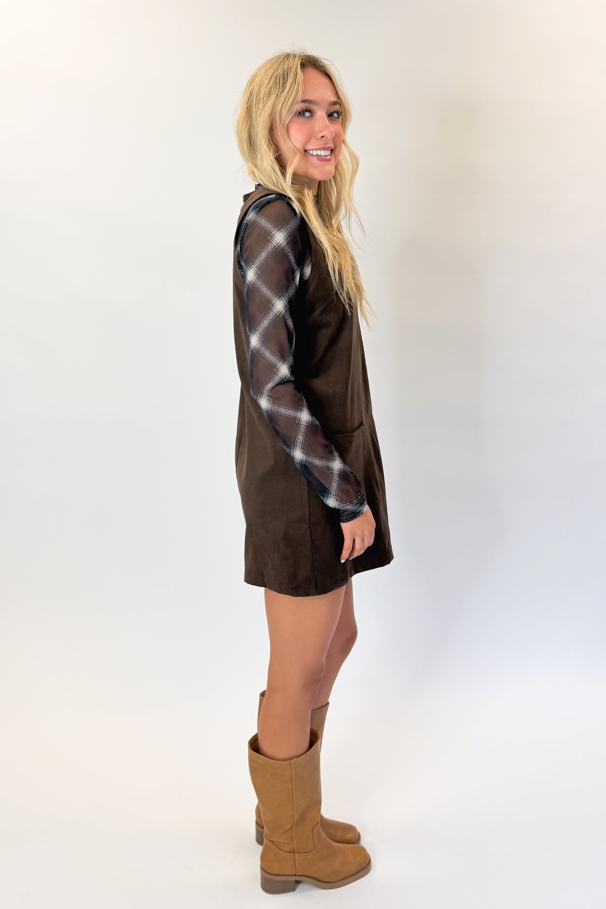 Side view of woman wearing Swagger Boutique’s Brown Suede V-Neck Mini Dress with Navy Plaid Mesh Top against white studio background.