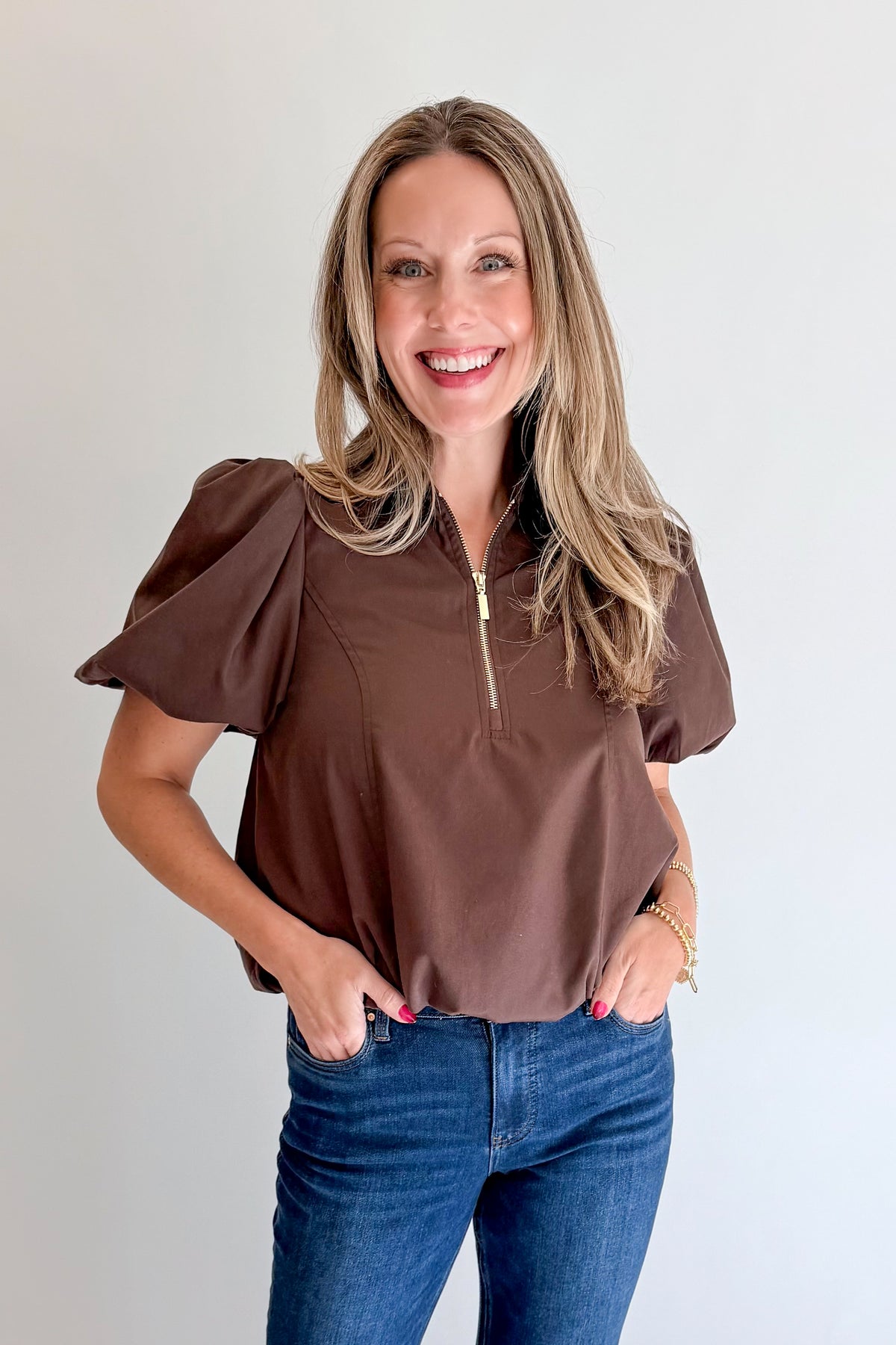 Front view of woman wearing Swagger Boutique’s Brown Zippered Bubble Hem Top by Entro with jeans against white studio background.