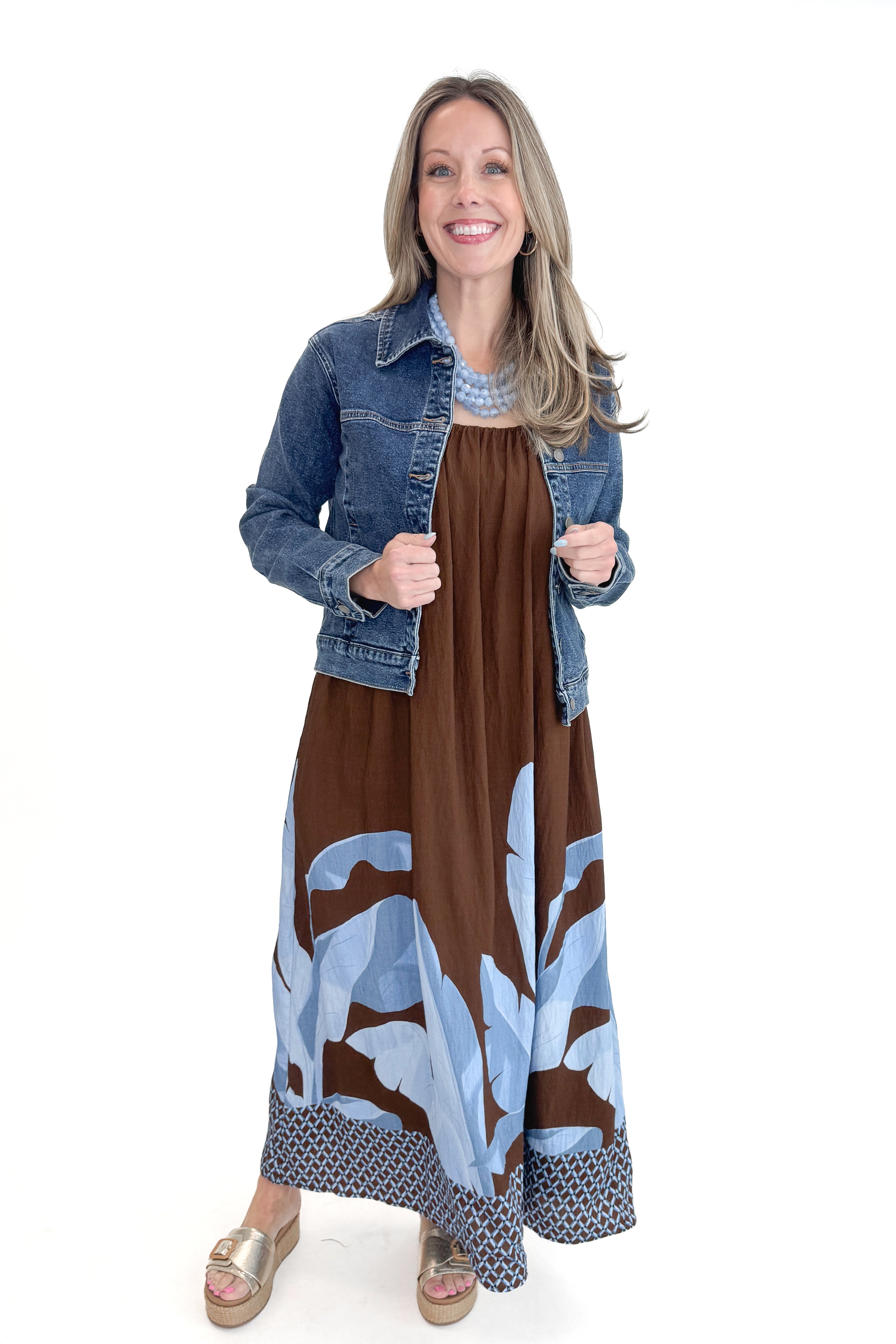 Front view of woman wearing Brown & Blue Floral Maxi Dress with gold sandals and Dark Wash Denim Jacket against white studio background.