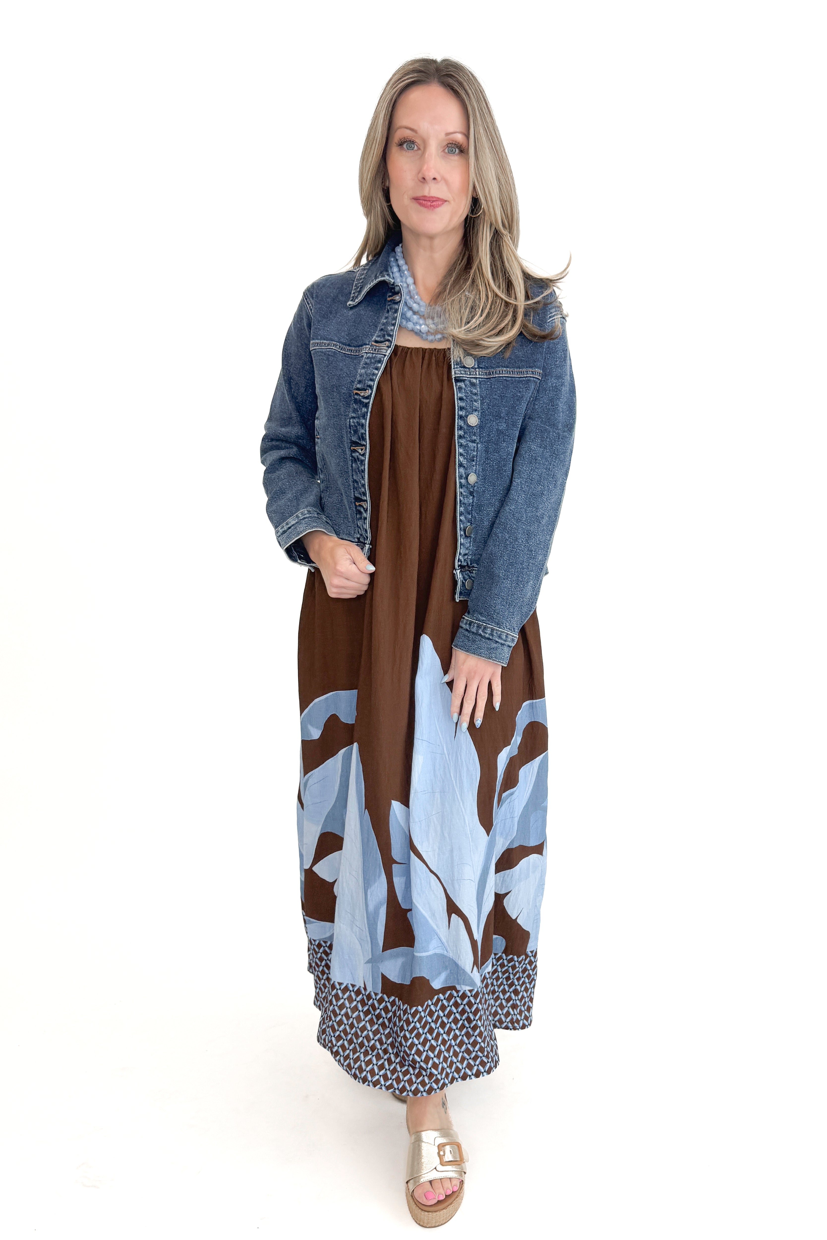 Front view of woman wearing Brown & Blue Floral Maxi Dress with gold sandals and Dark Wash Denim Jacket against white studio background.