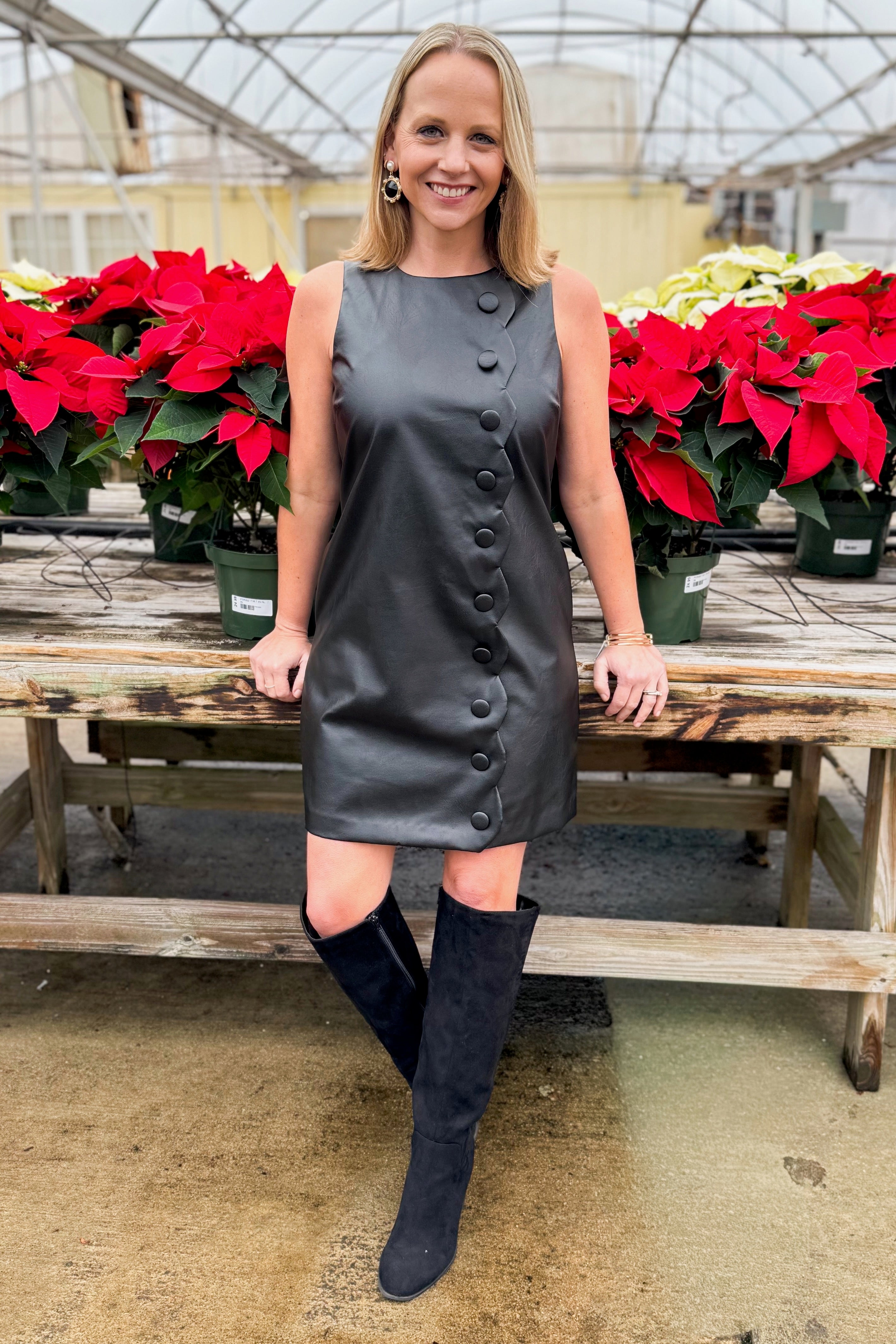 Front view of woman wearing Black Faux Leather Scallop Detail Dress with tall boots against around holiday decor.