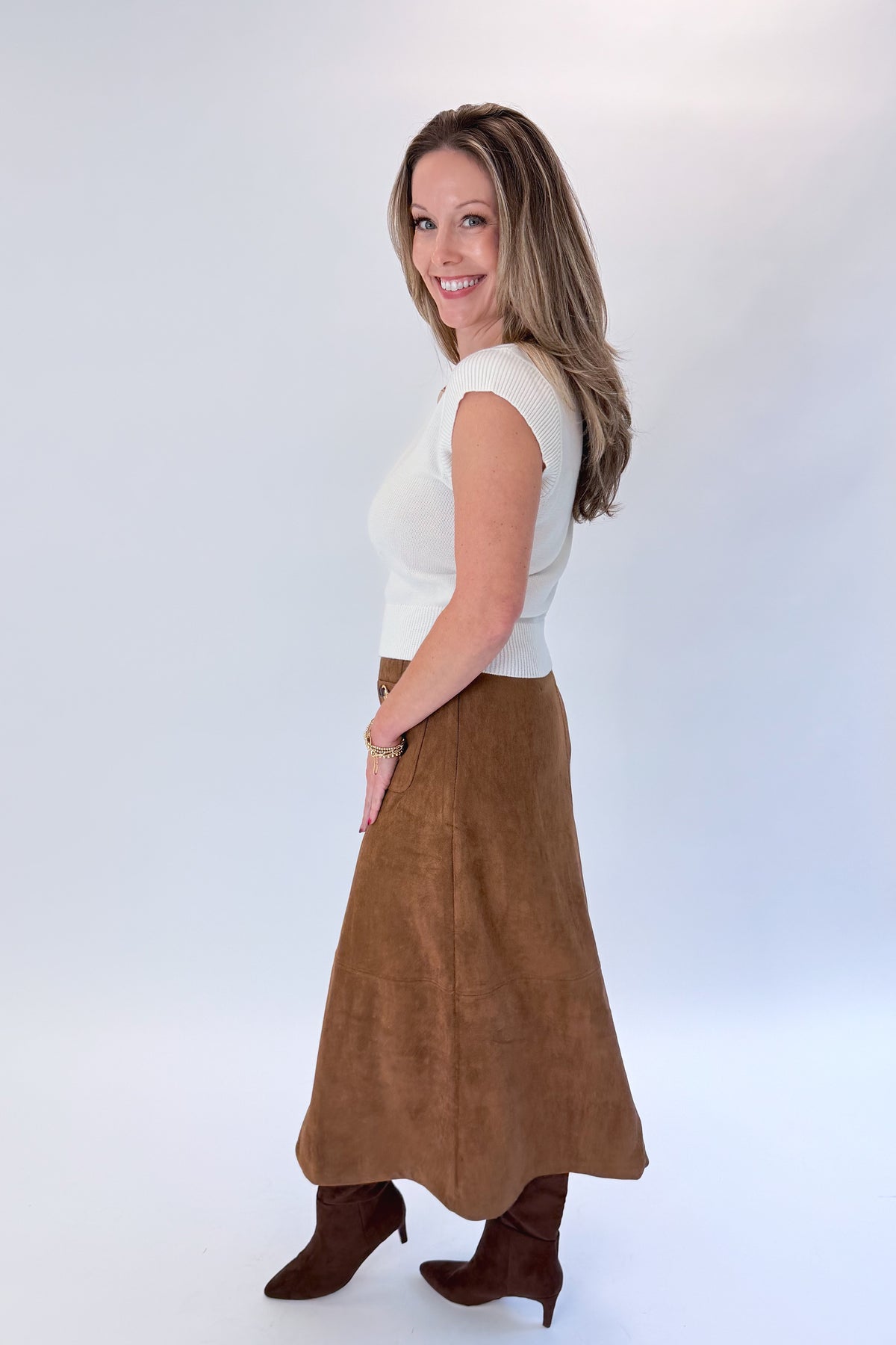 Side view of woman wearing Swagger Boutique’s White Cap Sleeve Sweater by PINCH with Camel Suede Midi Skirt With Pockets by Entro against white studio background.