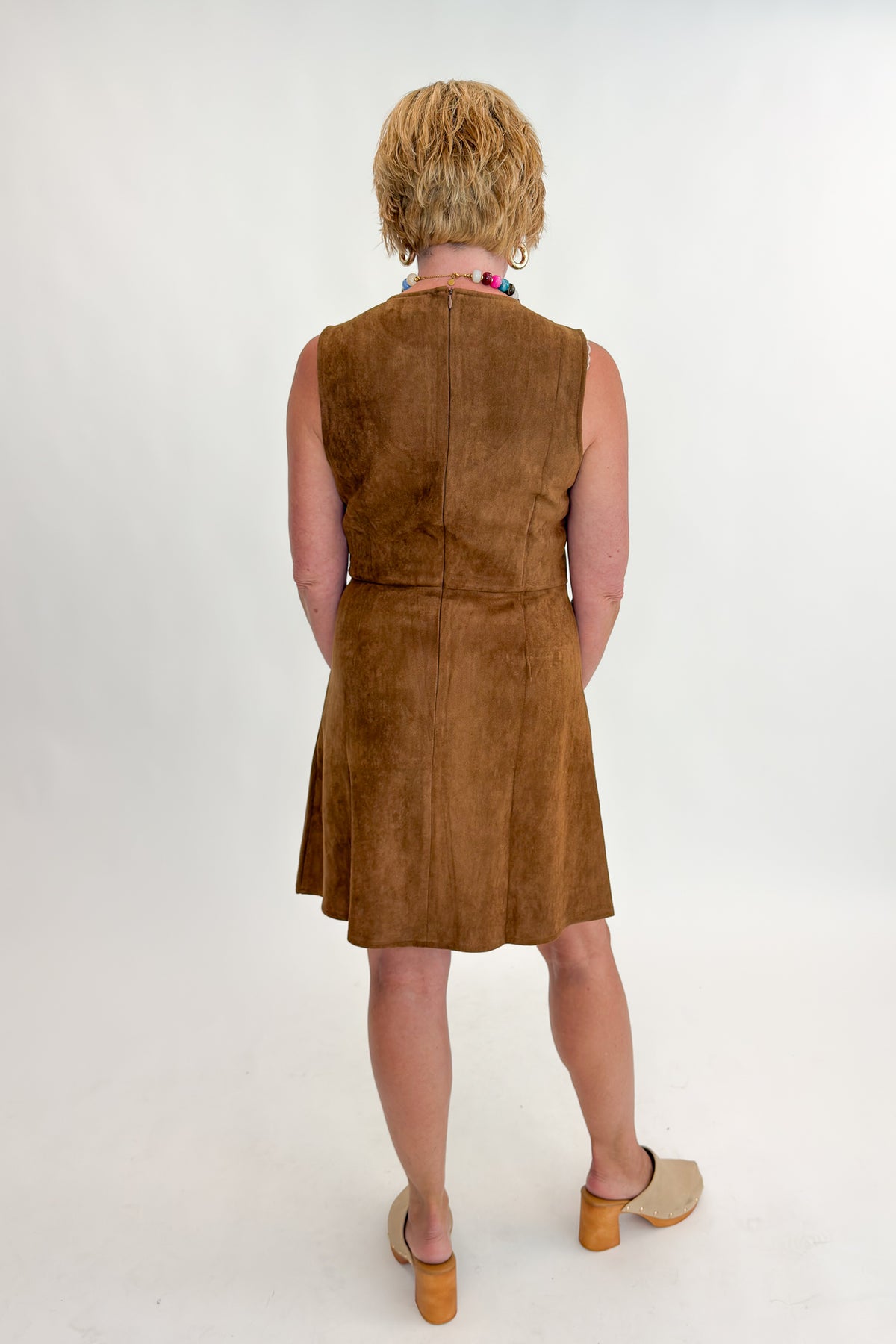 Back view of woman wearing Swagger Boutique’s Camel Suede Mini Dress With Pockets by Entro with clogs against white studio background.