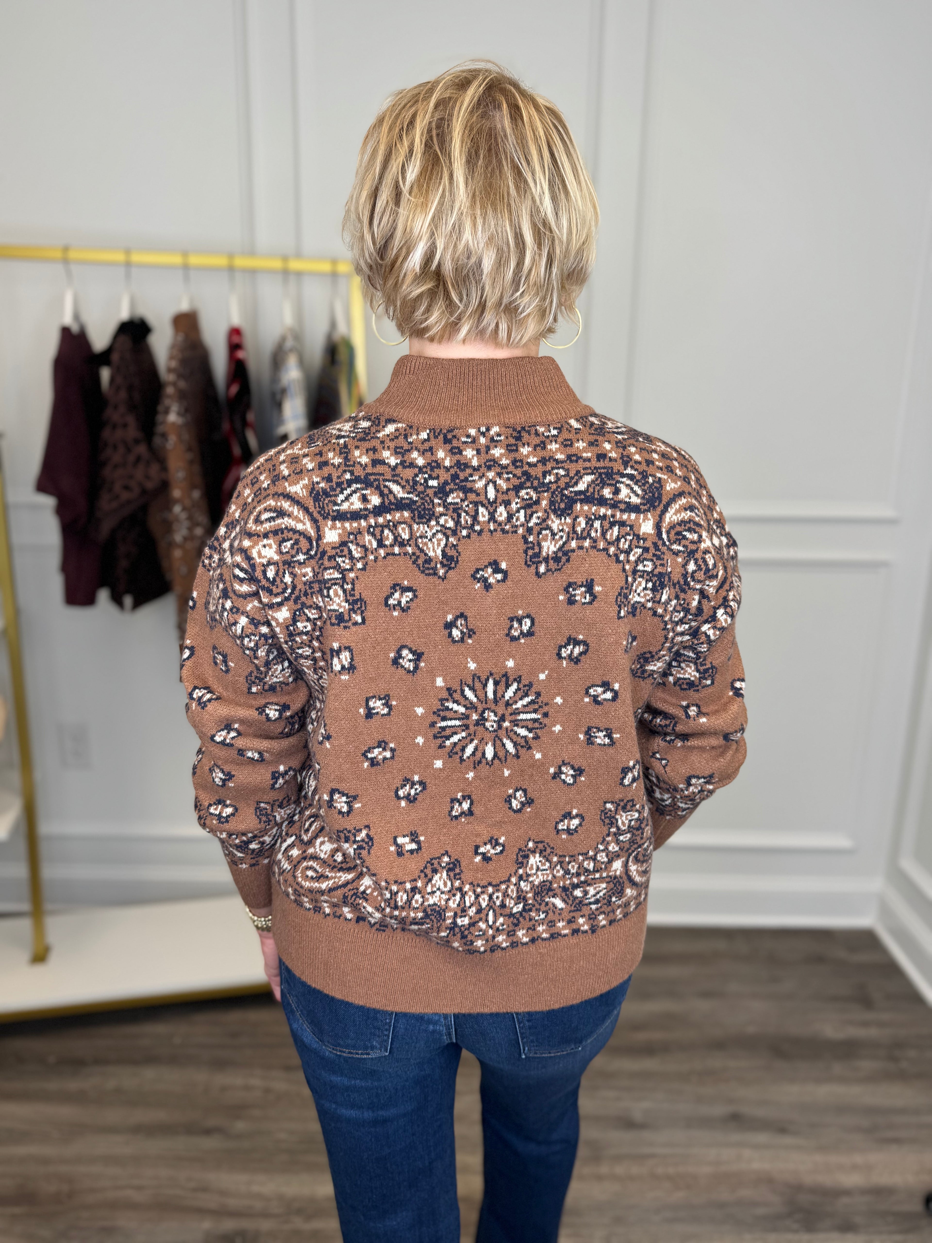 Back view of woman wearing Swagger Boutique’s Camel Bandana Print Half Zip Sweater with jeans with clothing hanging in the background.