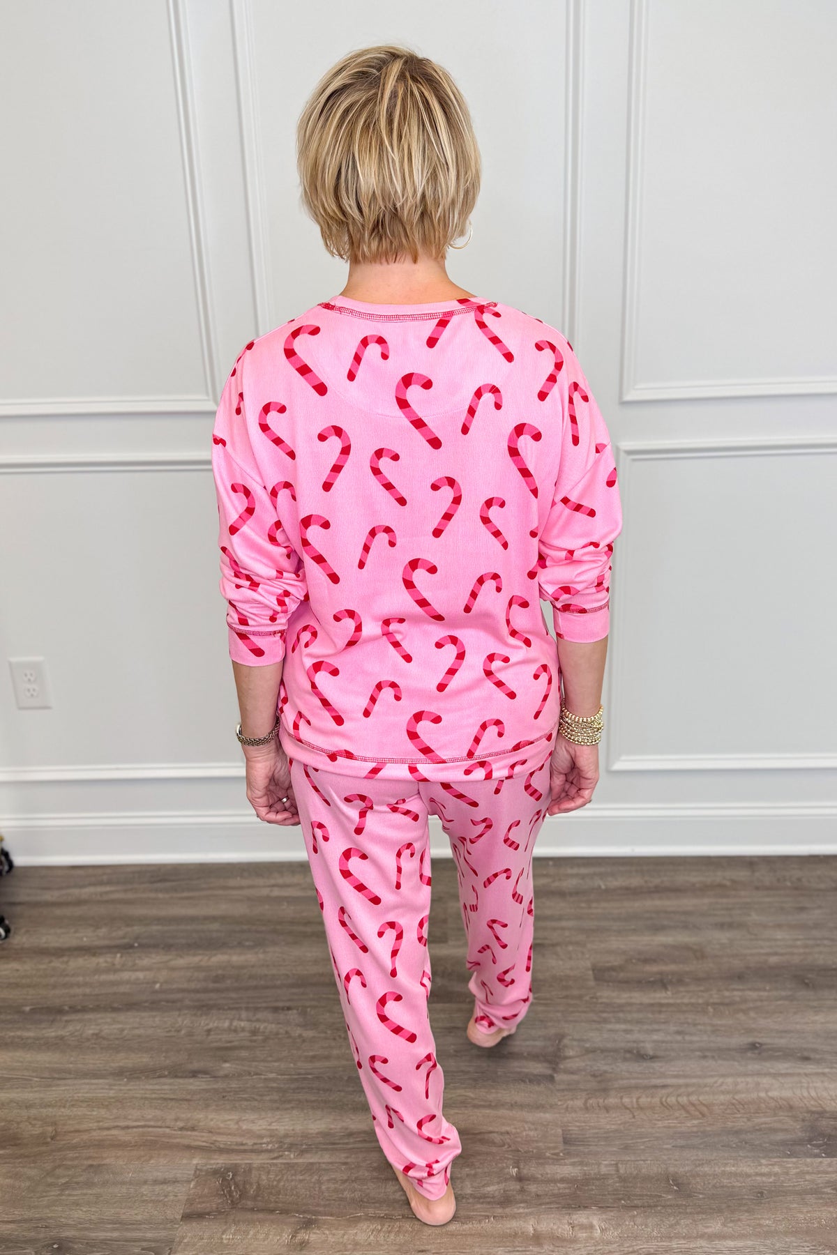 A woman viewed from behind wearing a pink long-sleeve pajama top and matching pants, both covered in a red candy cane pattern, standing on a wood floor against a white paneled wall.