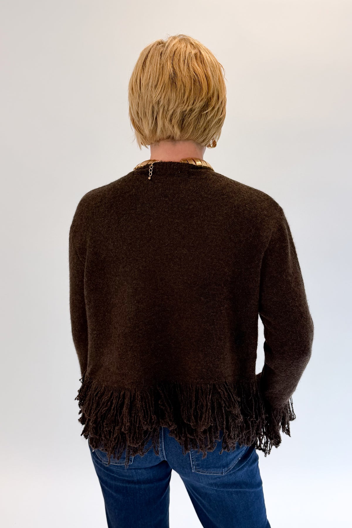 A person viewed from the back wearing a dark brown fuzzy knit sweater with fringe trim at the hem and cuffs, paired with blue jeans, against a plain light gray studio background.