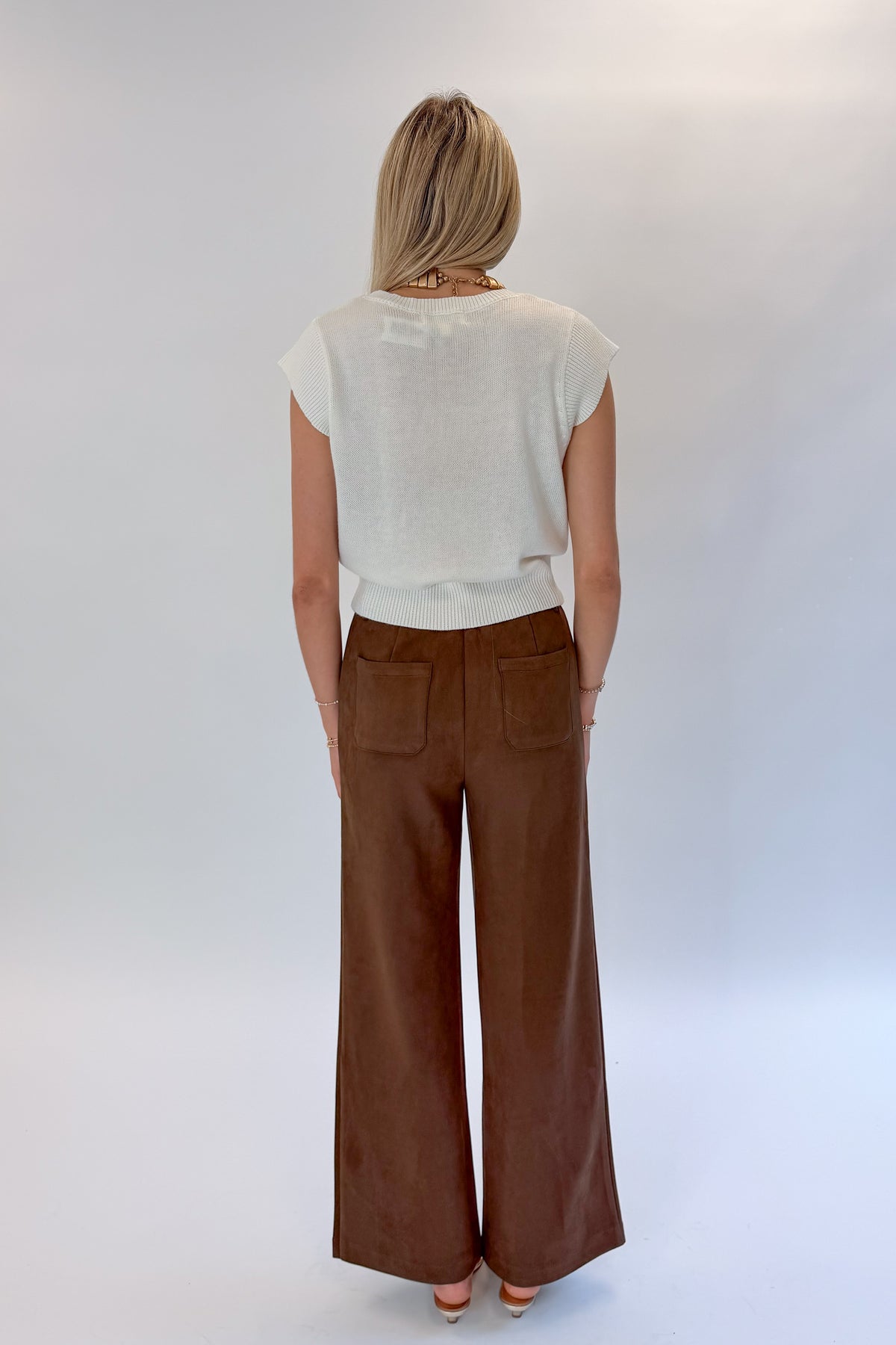 Back view of woman wearing Swagger Boutique’s Chocolate Brown Suede Pants with Living My Best Life White Cap Sleeve Sweater by PINCH against white studio background.