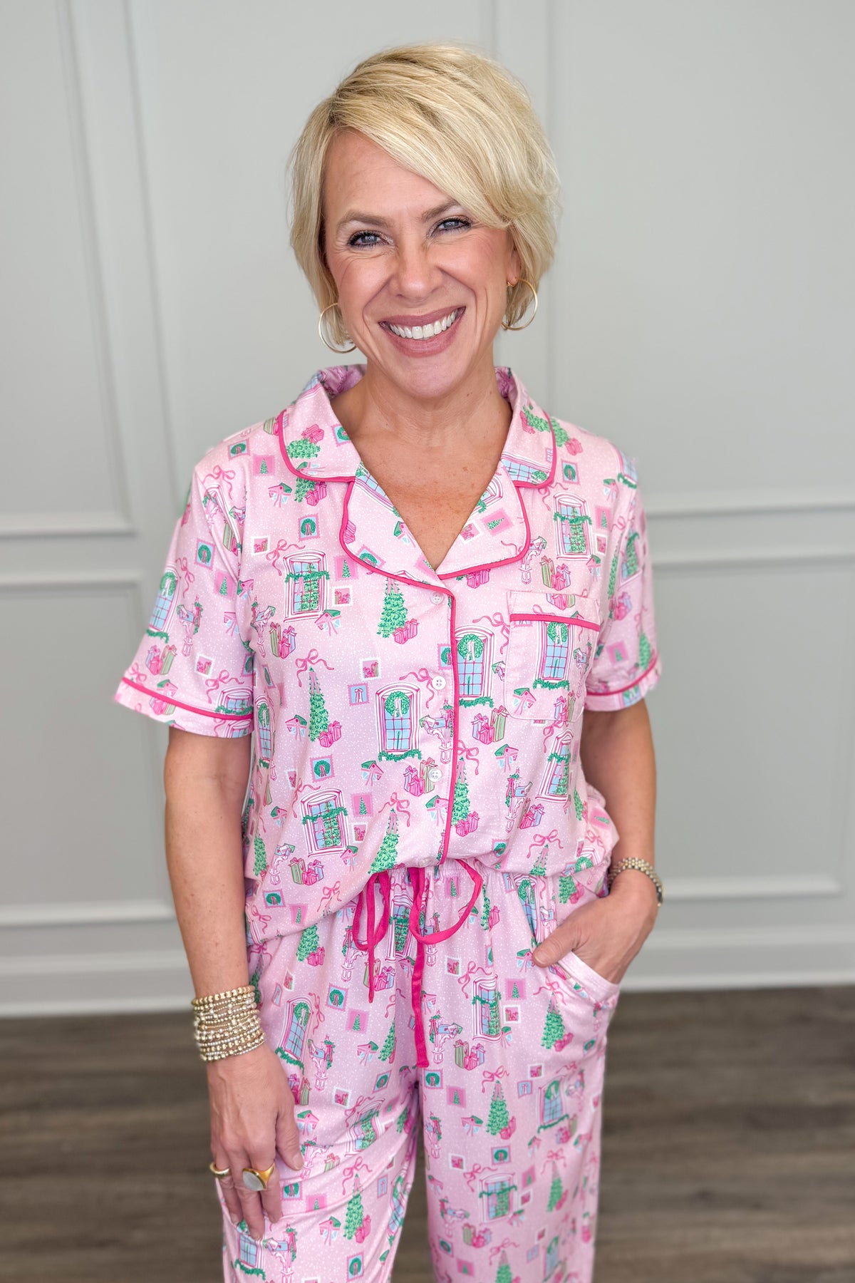 Model wearing the Christmas Cottage Charlotte Pajama Set in pink, featuring a festive Christmas cottage print with green trees, holiday bows, and presents. The set includes a short-sleeve button-down top with contrast piping and matching long pajama pants.