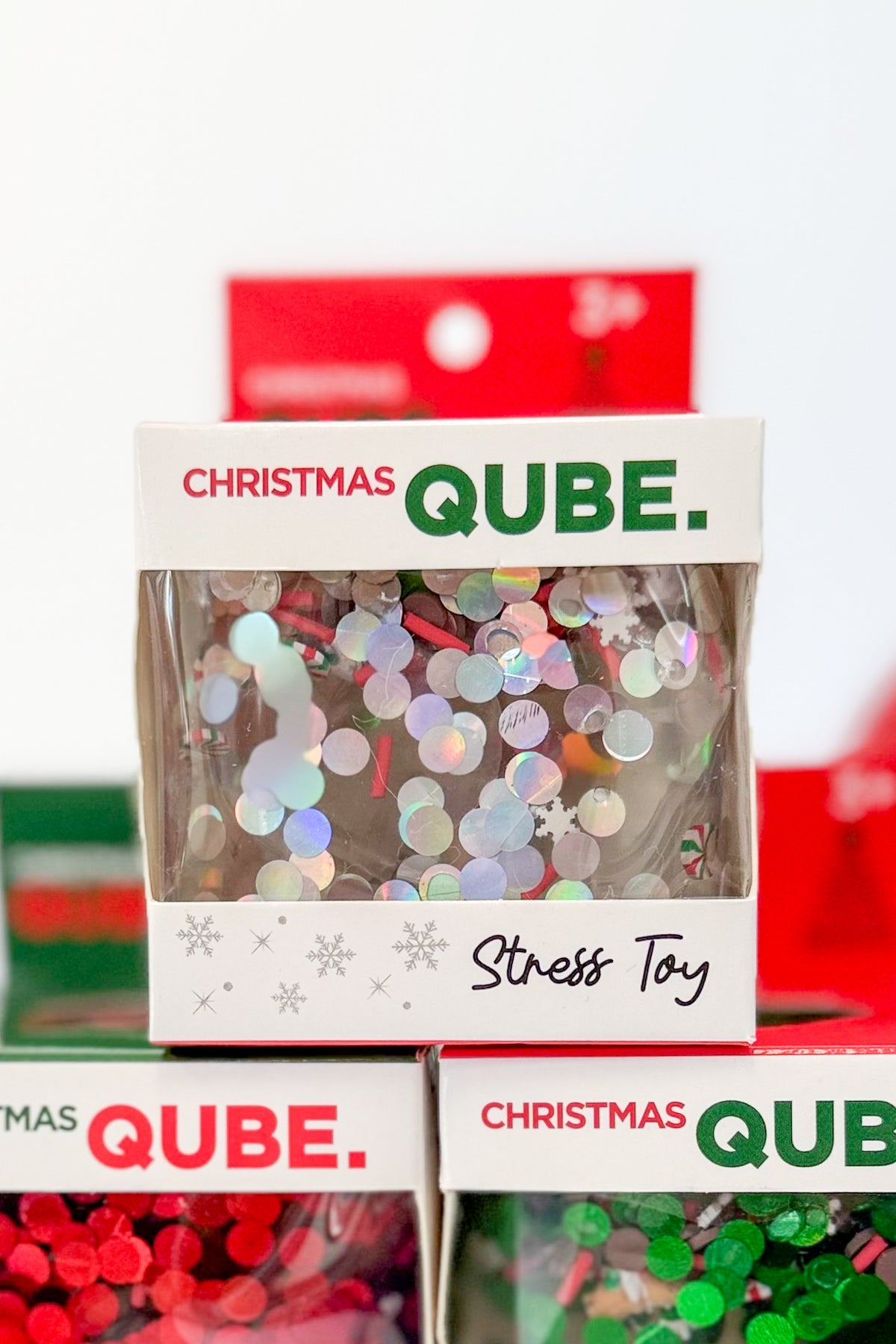 A stack of small retail boxes labeled Christmas Qube. Stress Toy, showing iridescent round confetti pieces visible through clear front windows.