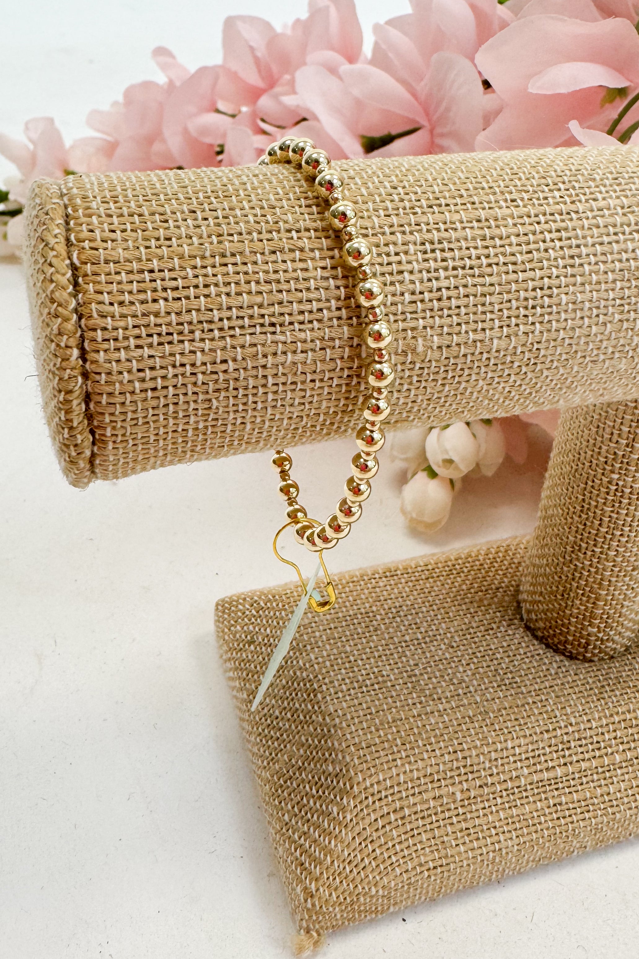 Gold beaded bracelet on a beige jewelry display with pink flowers in the background