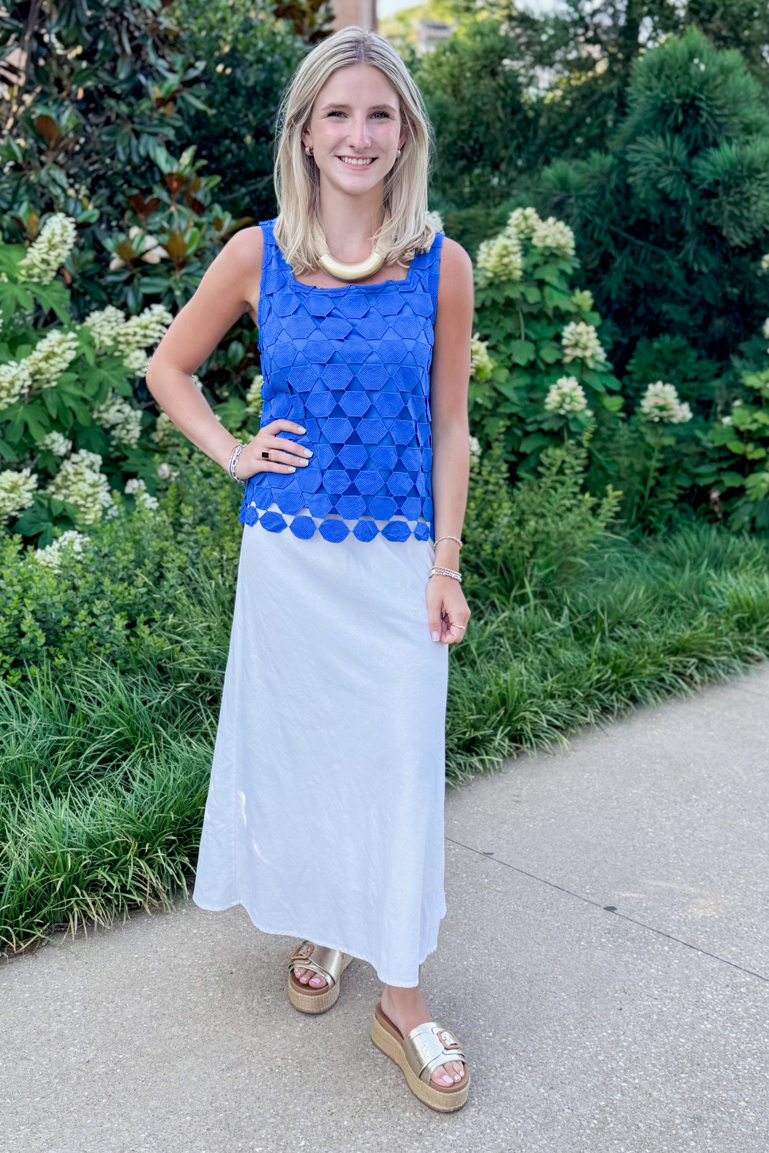 Front view of woman wearing Swagger Boutiqueās Cobalt Sleeveless Lace Tank with white skirt standing outside by trees.