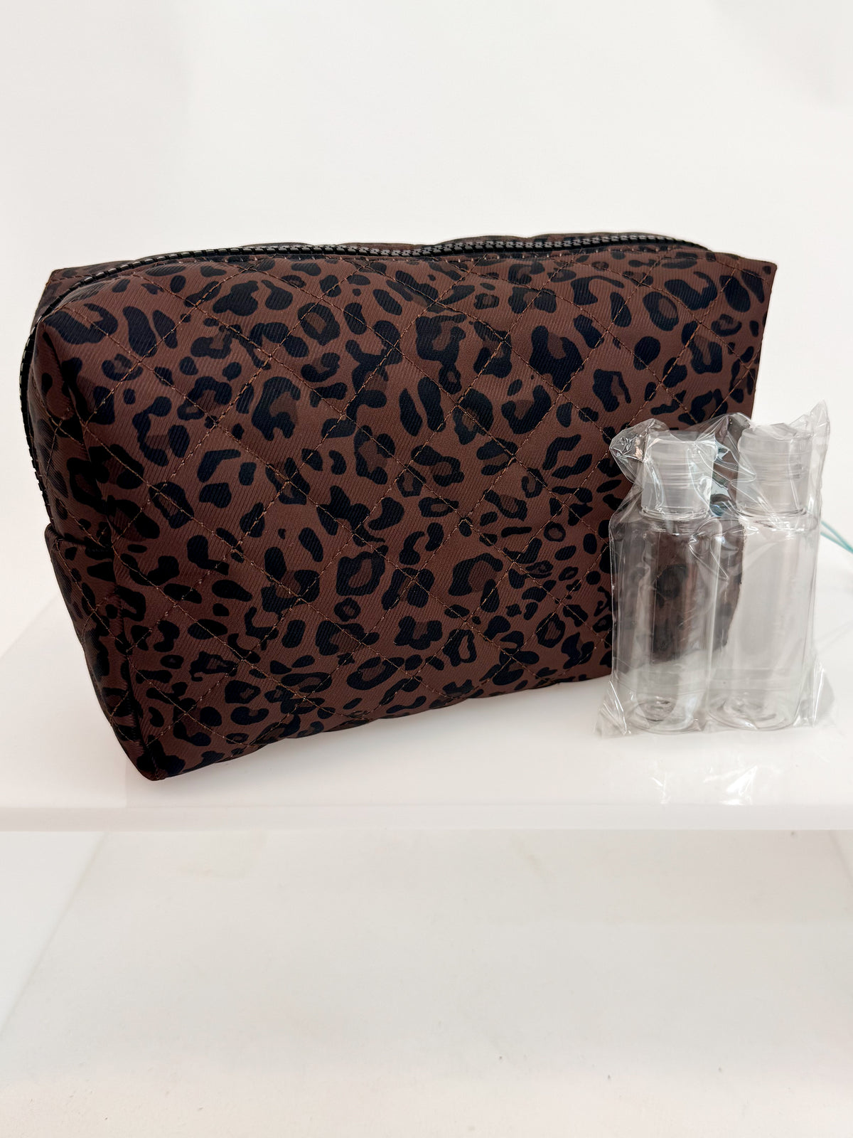 A brown and black leopard print zippered cosmetic pouch displayed next to two small clear plastic travel bottles on a white surface.