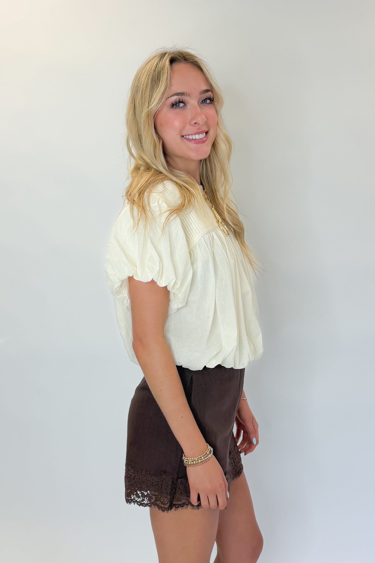 Side view of woman wearing Swagger Boutique’s Brown Lace Lined Denim Skort with Cream Mixed Material Bubble Hem Top against white studio background.