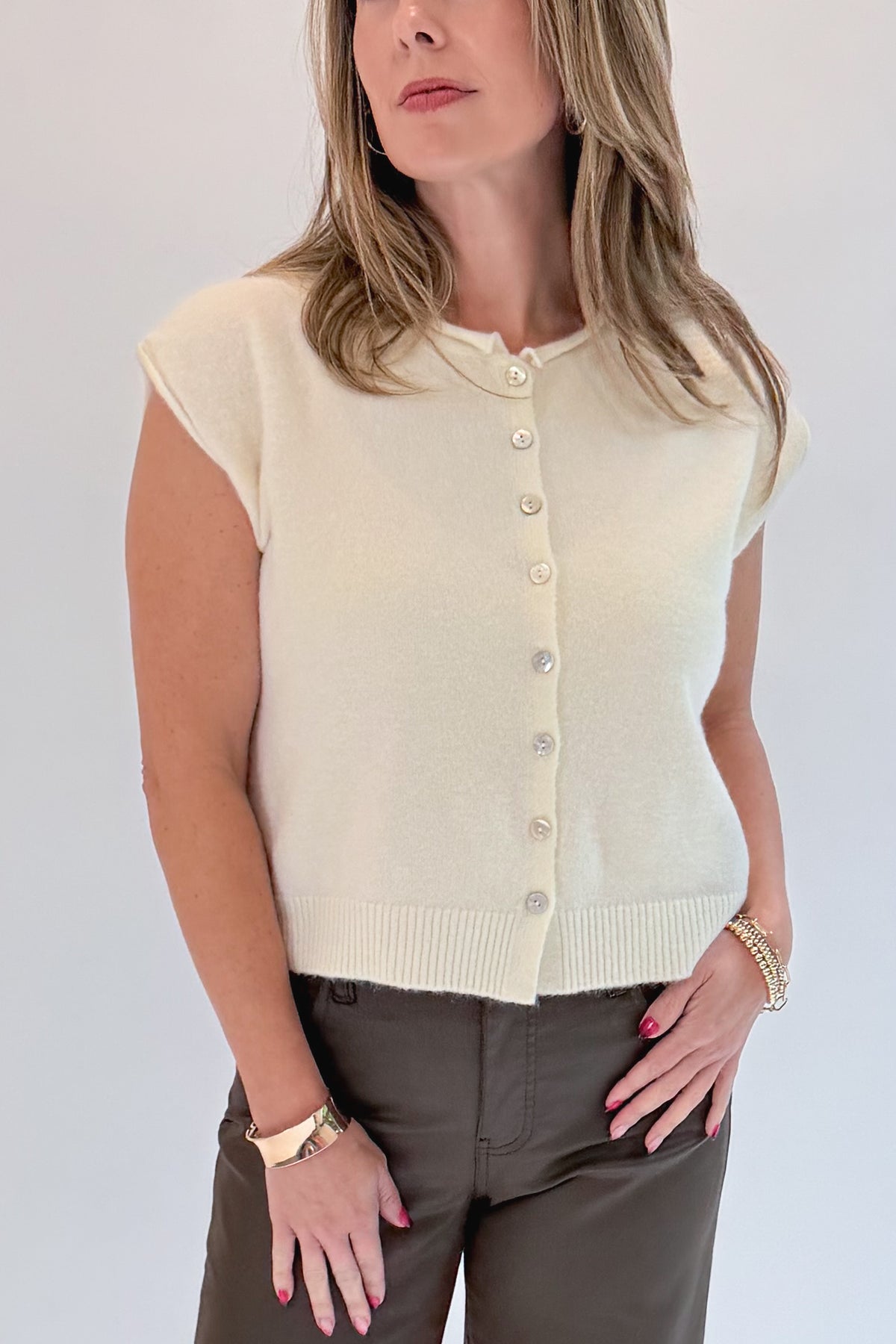 A woman modeling a cream-colored short-sleeve knit top with a button-up front, paired with dark brown pants, against a plain light gray background.