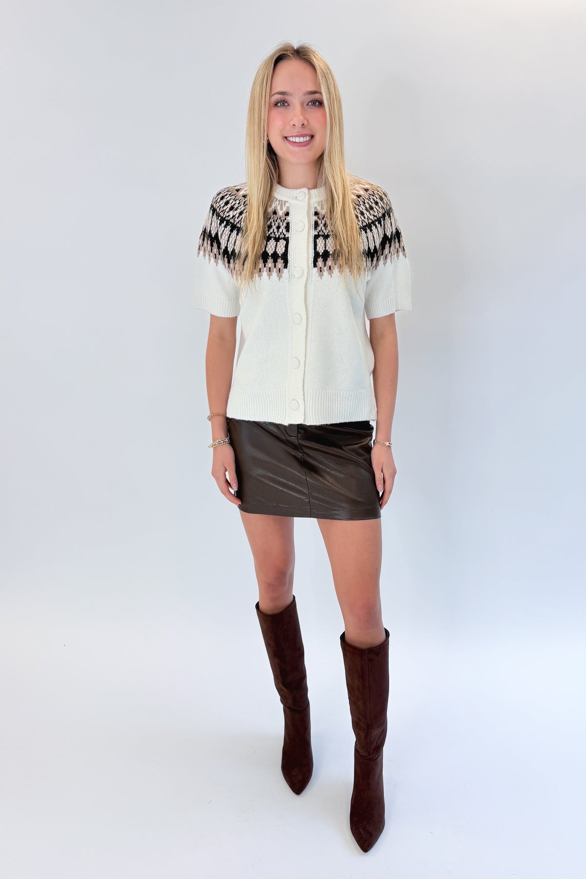 Front view of woman wearing Swagger Boutique’s Cream & Brown Fair Isle Button Up Short Sleeve Sweater with brown faux leather mini skirt against white studio background.