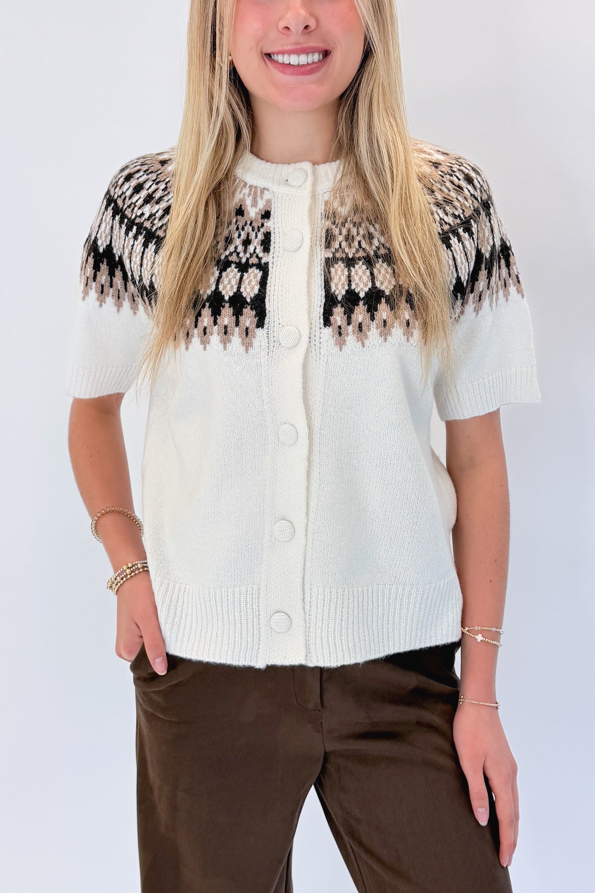 Front detail view of woman wearing Swagger Boutique’s Cream & Brown Fair Isle Button Up Short Sleeve Sweater with Chocolate Brown Suede Pants against white studio background.