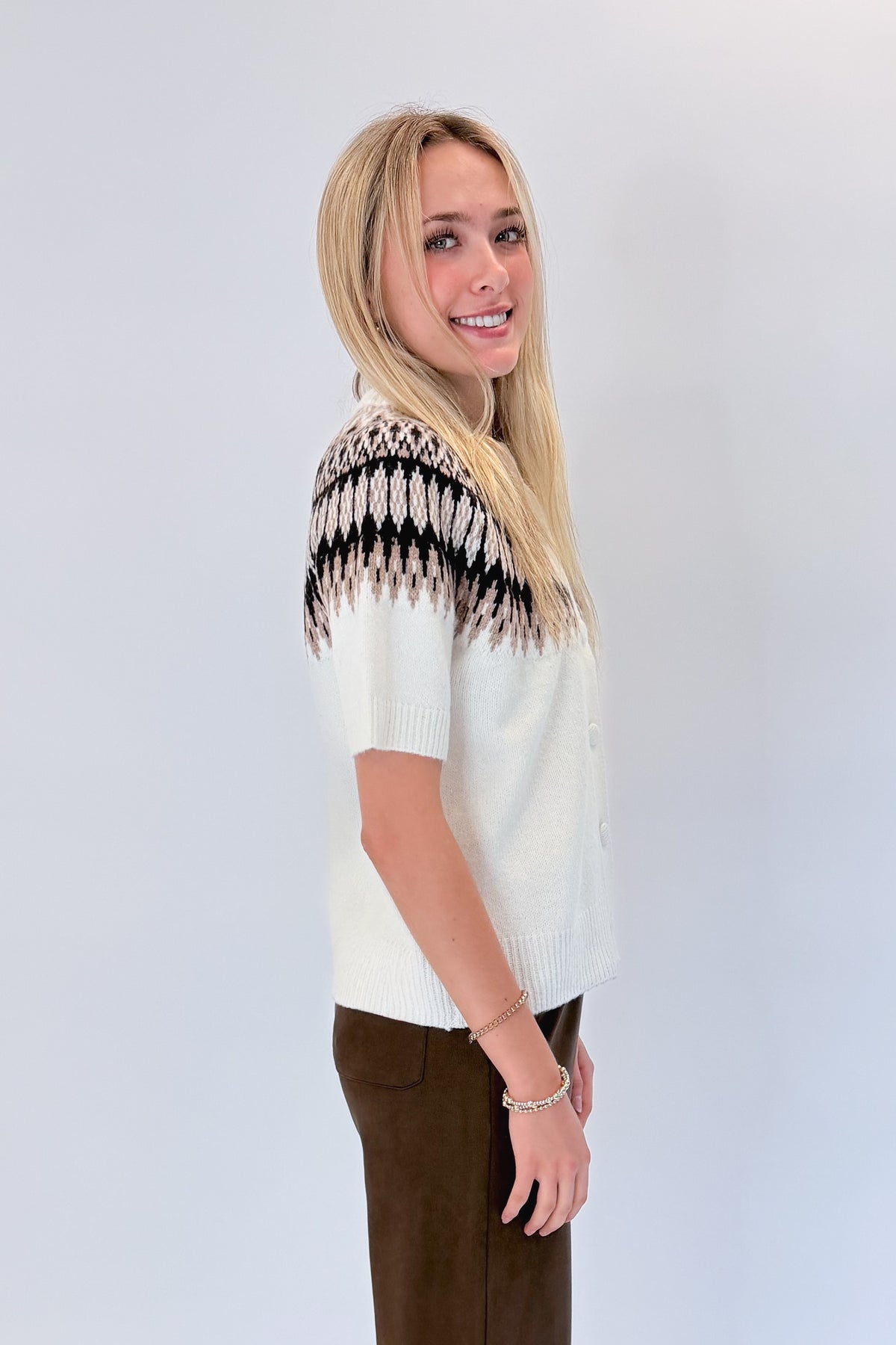 Side view of woman wearing Swagger Boutique’s Cream & Brown Fair Isle Button Up Short Sleeve Sweater with Chocolate Brown Suede Pants against white studio background.