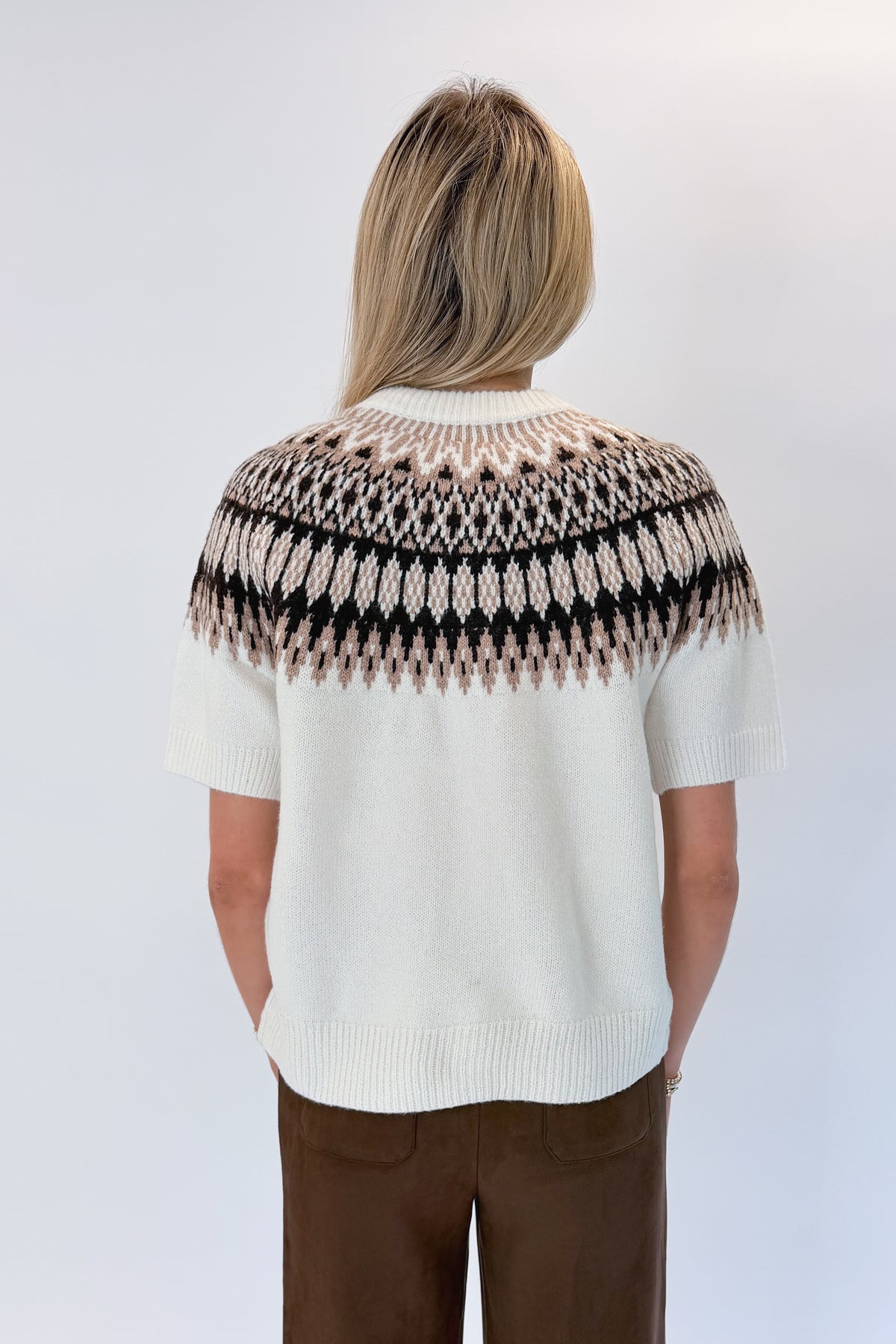 Back view of woman wearing Swagger Boutique’s Cream & Brown Fair Isle Button Up Short Sleeve Sweater with Chocolate Brown Suede Pants against white studio background.