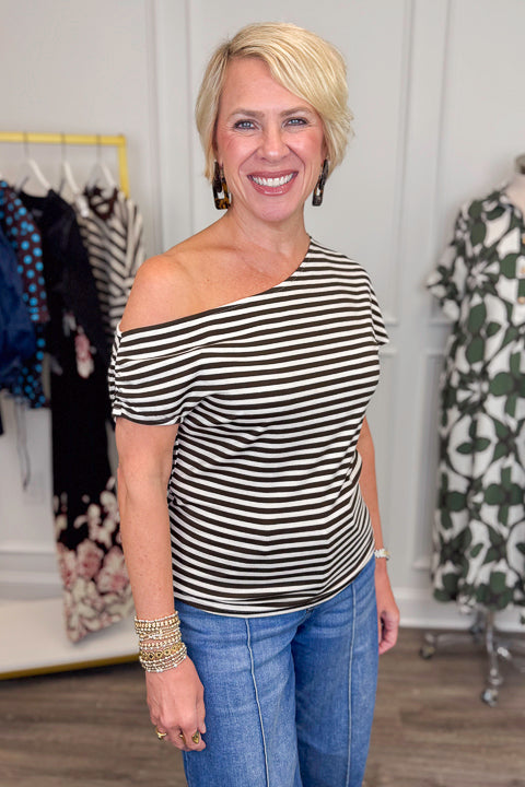 Front view of woman wearing Swagger Boutiqueās Cream & Brown Striped Asymmetrical Top with jeans with clothing hanging in the background.