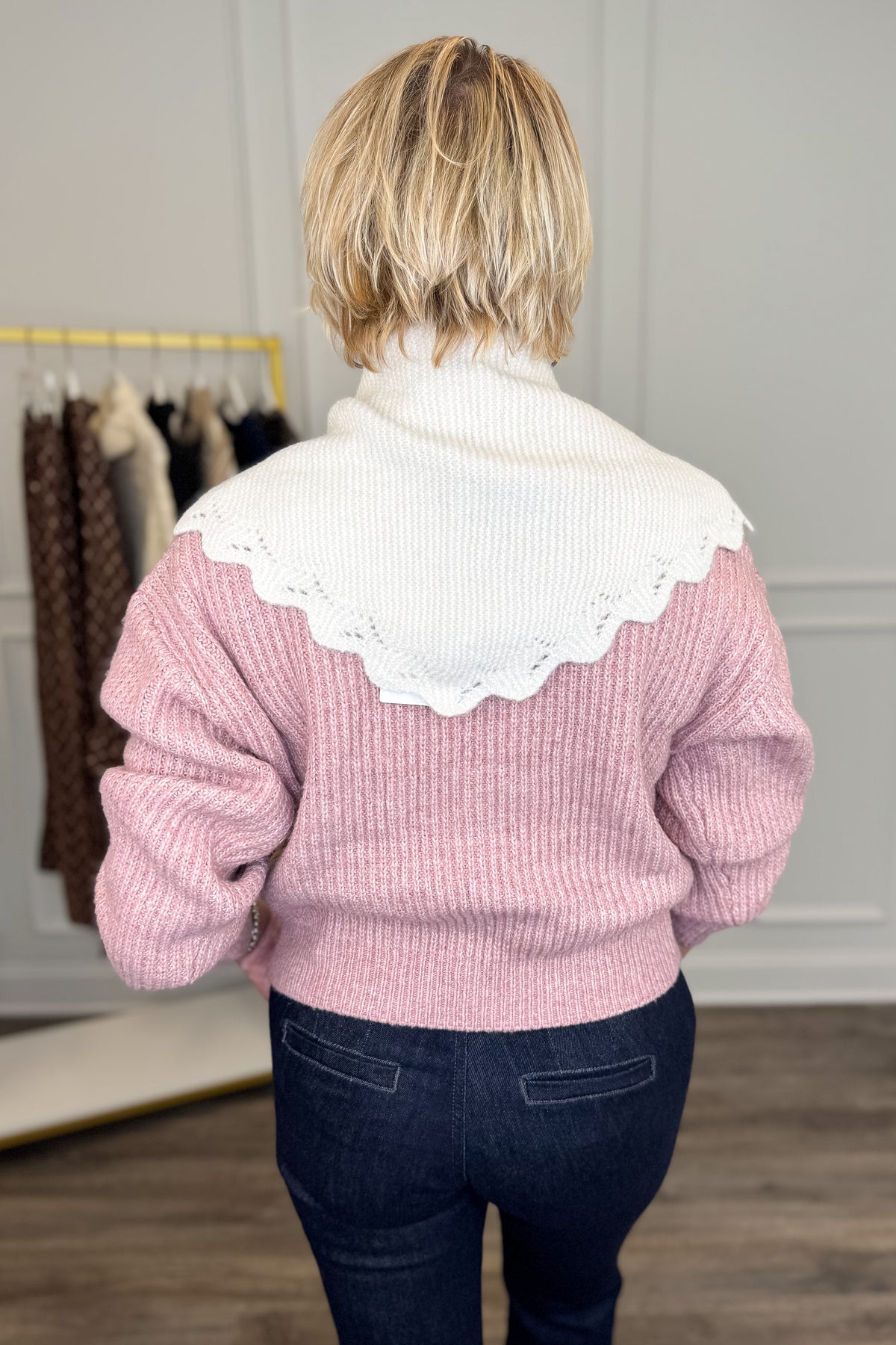 A woman viewed from behind wearing a pink ribbed knit sweater with a large white scalloped knit scarf paired with dark blue jeans in an indoor boutique setting.