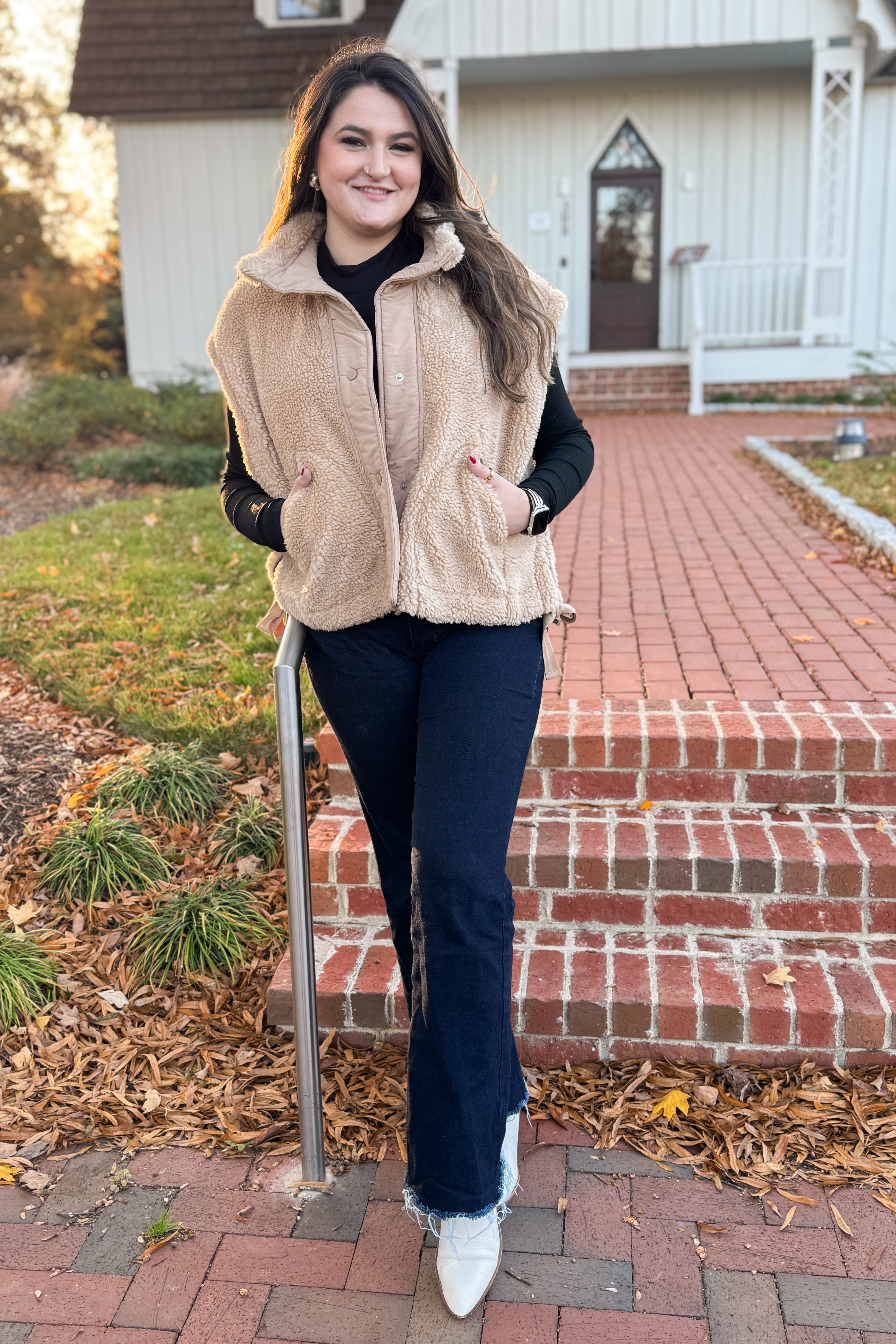 Woman wearing a beige vest over a black top and blue jeans, standing on a brick path with a house in the background.