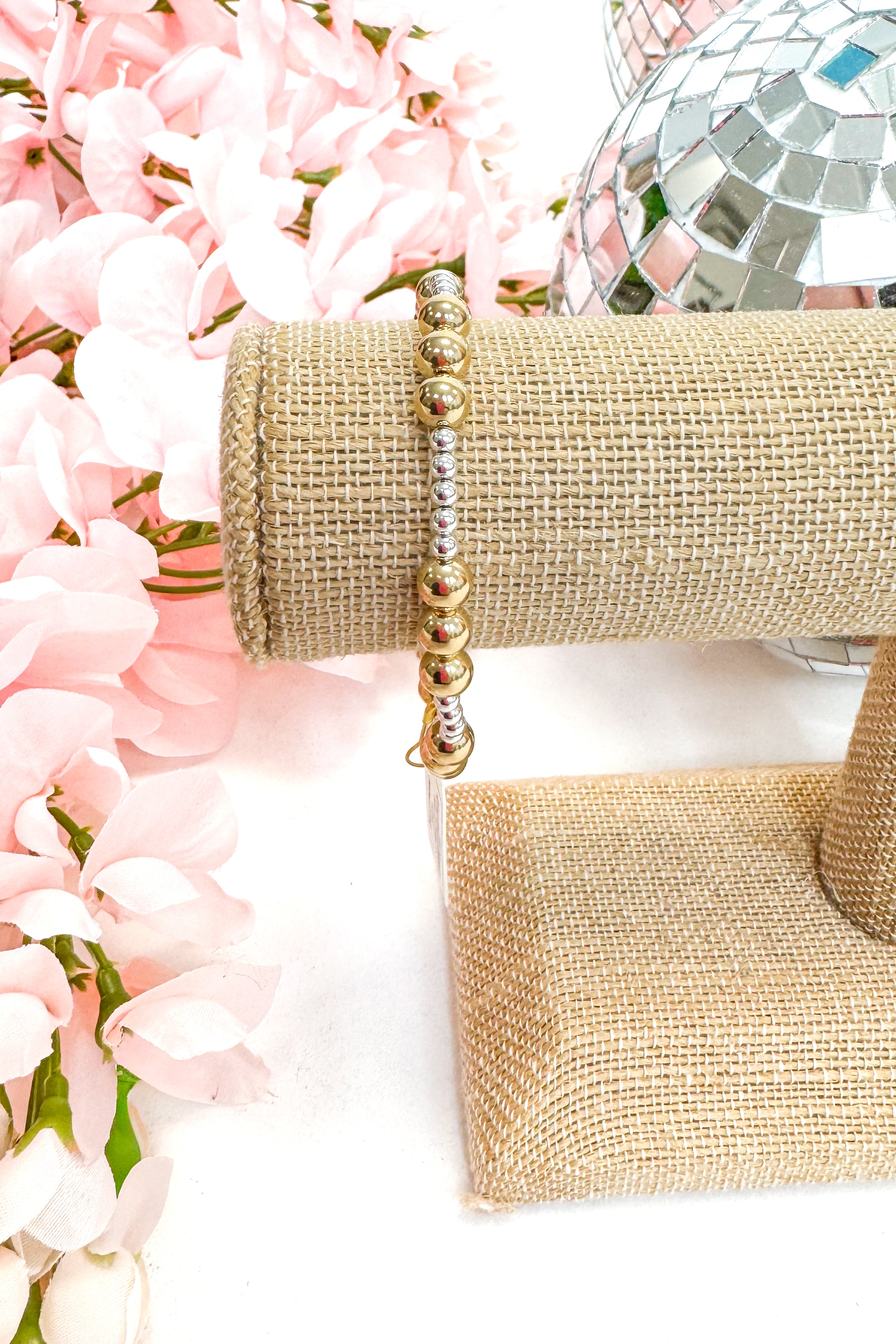 Gold beaded bracelet on a burlap roll with pink flowers and disco ball in the background