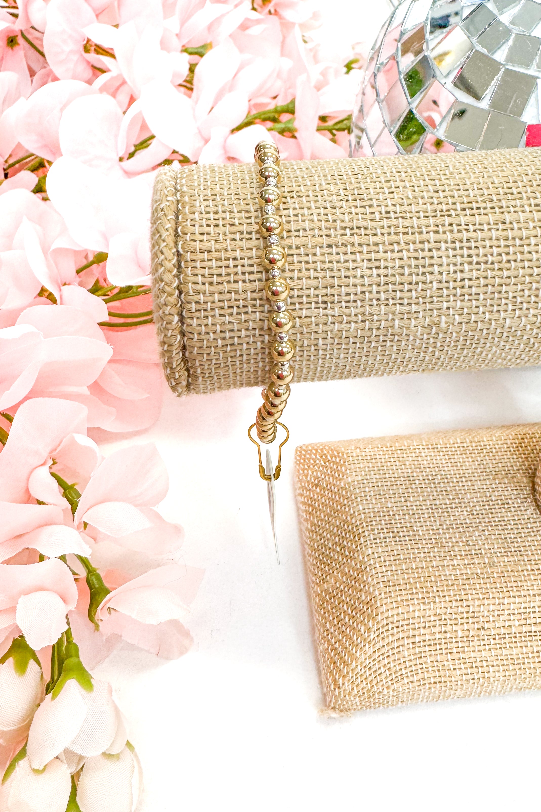 Gold beaded bracelet on a white surface with pink flowers in the background