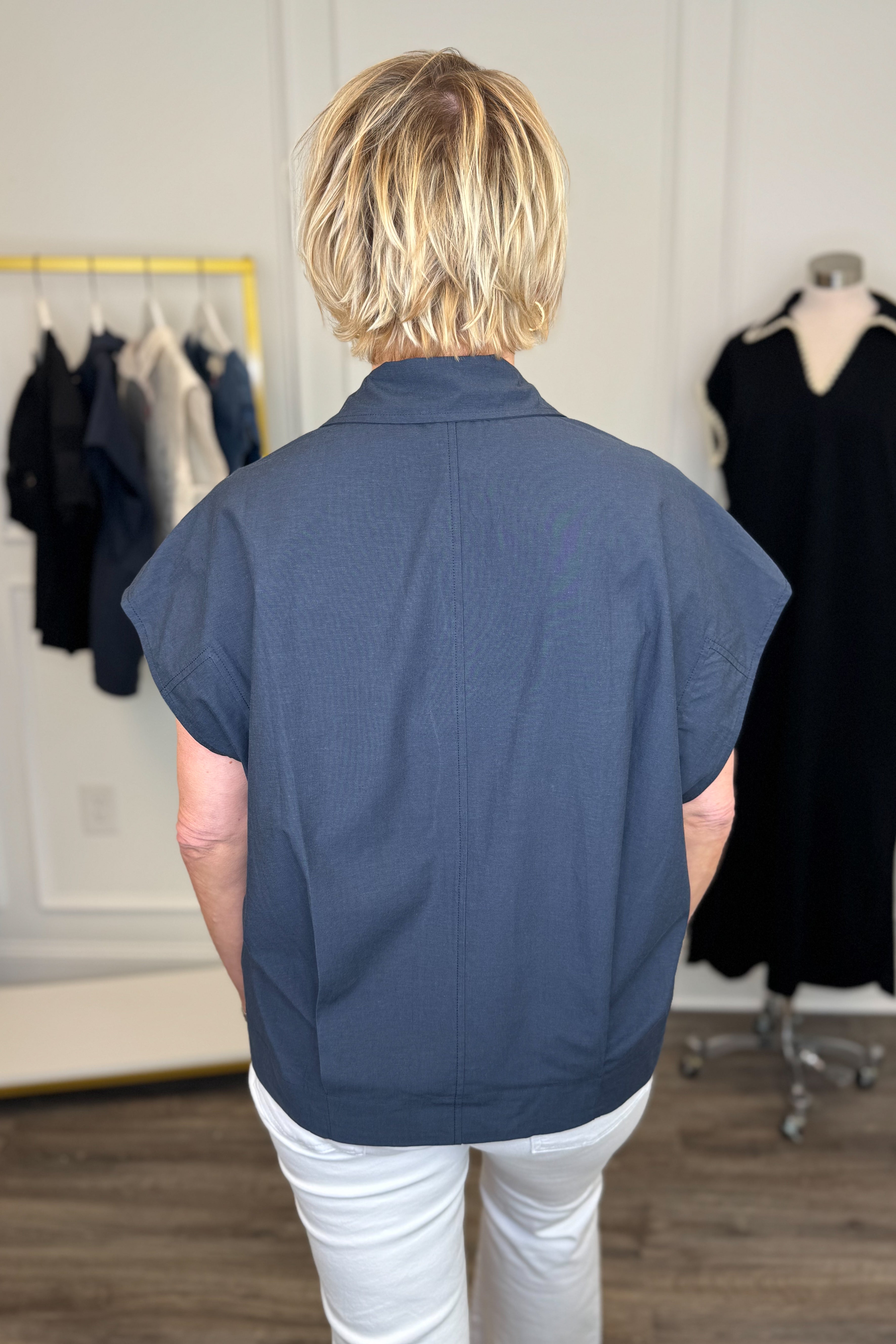 Back view of woman wearing Swagger Boutiqueβs Navy Collared Cap Sleeve Top with jeans with clothing hanging in the background.