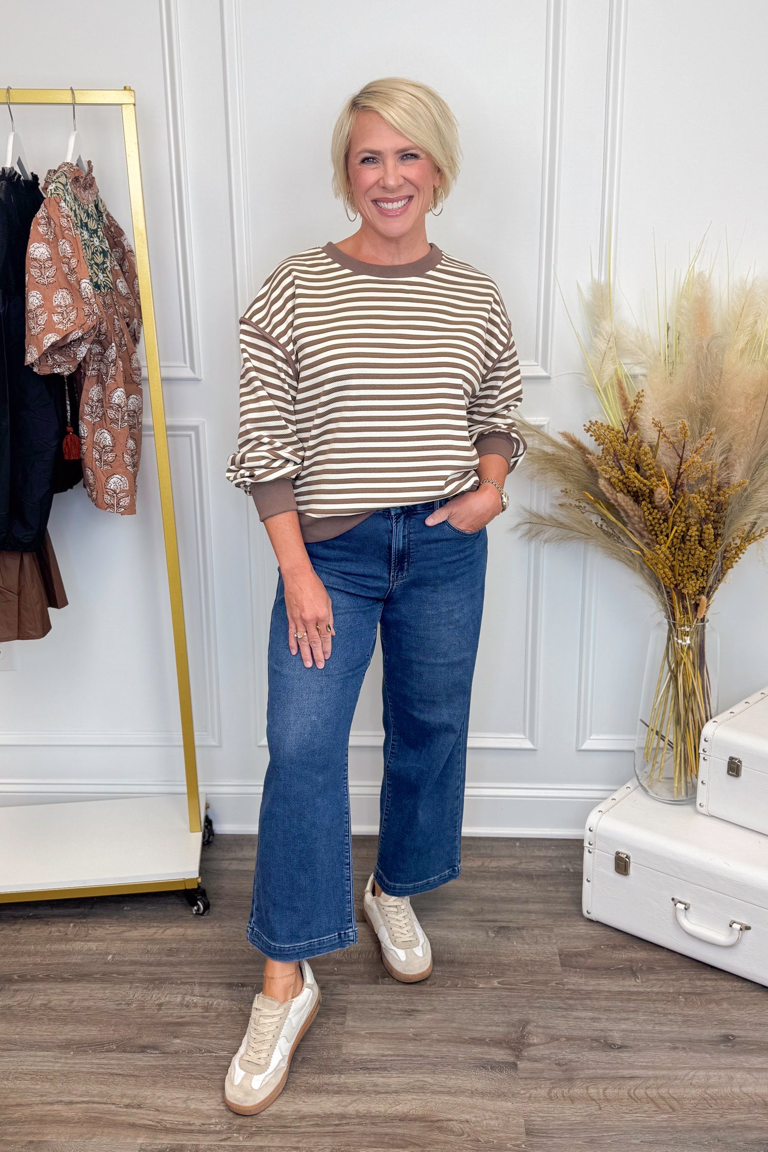 Woman wearing a striped sweater and blue jeans standing in a room with a coat rack and decorative plants.