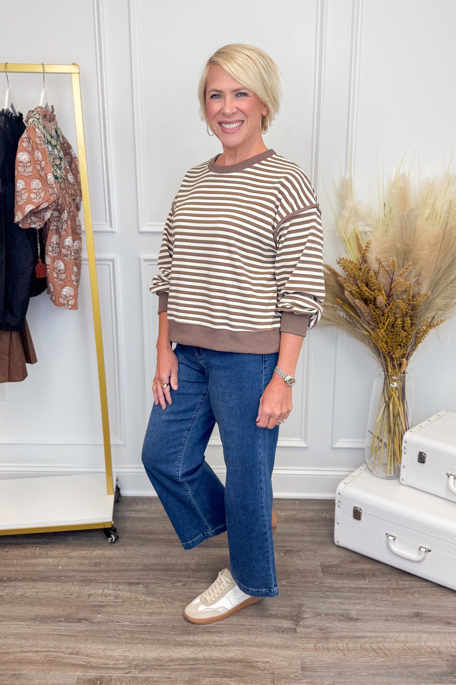 Woman wearing a striped sweater and blue jeans standing in a room with a white wall and wooden floor.
