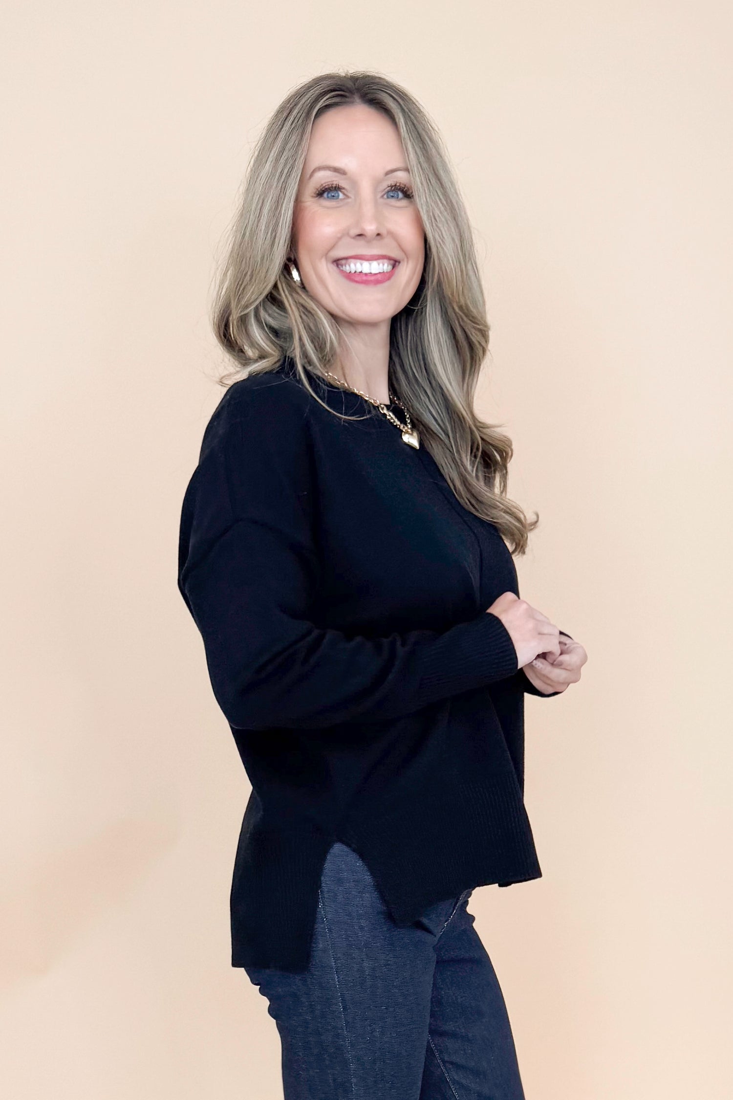 Side view of woman wearing black long sleeve sweater with jeans against tan studio background.