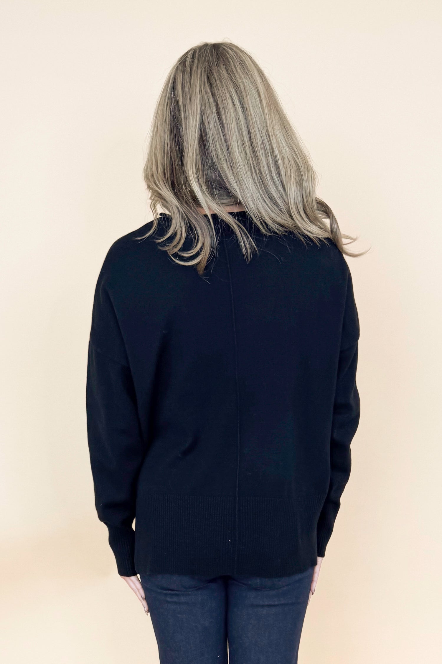 Back view of woman wearing black long sleeve sweater with jeans against tan studio background.