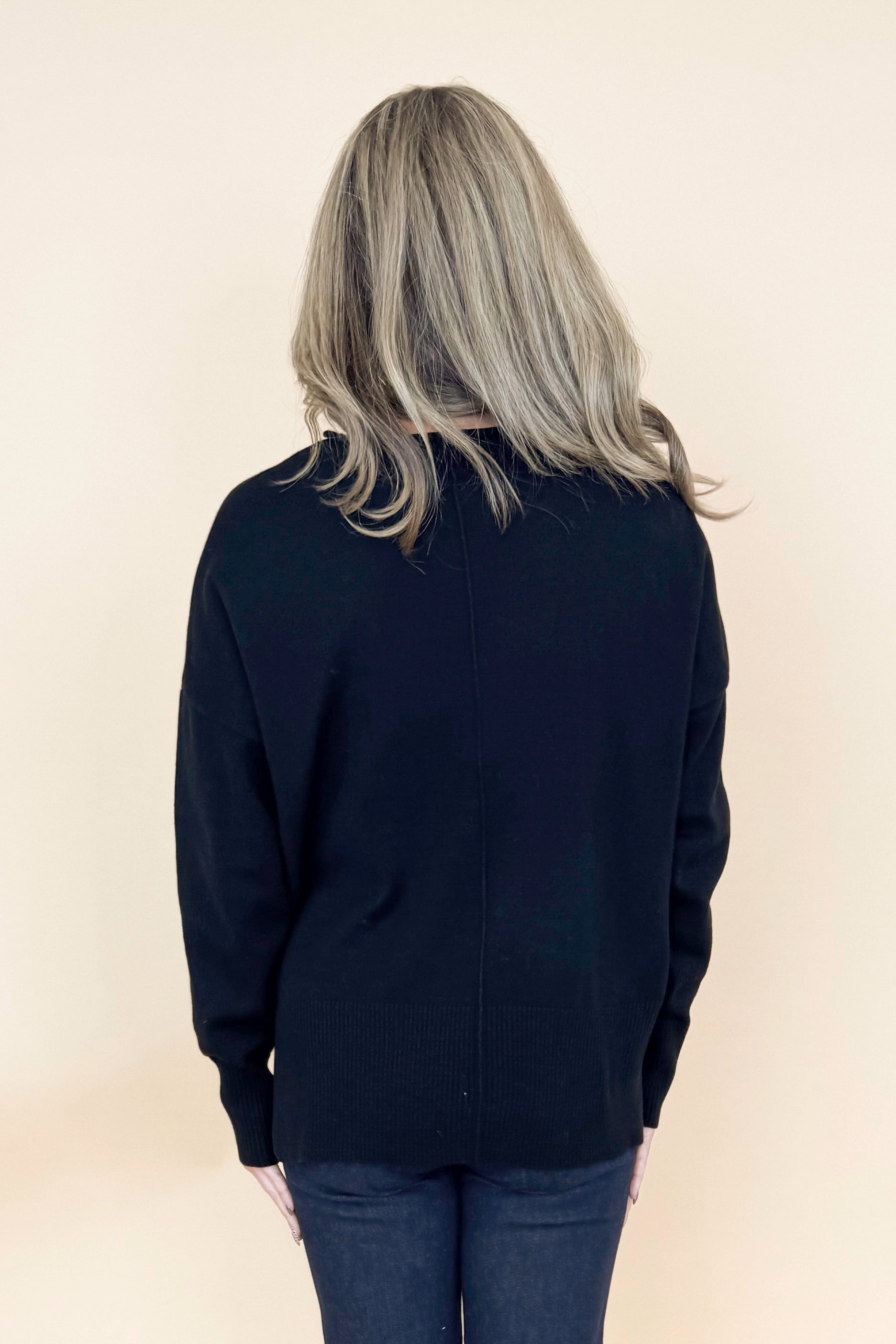 Back view of woman wearing black long sleeve sweater with jeans against tan studio background.