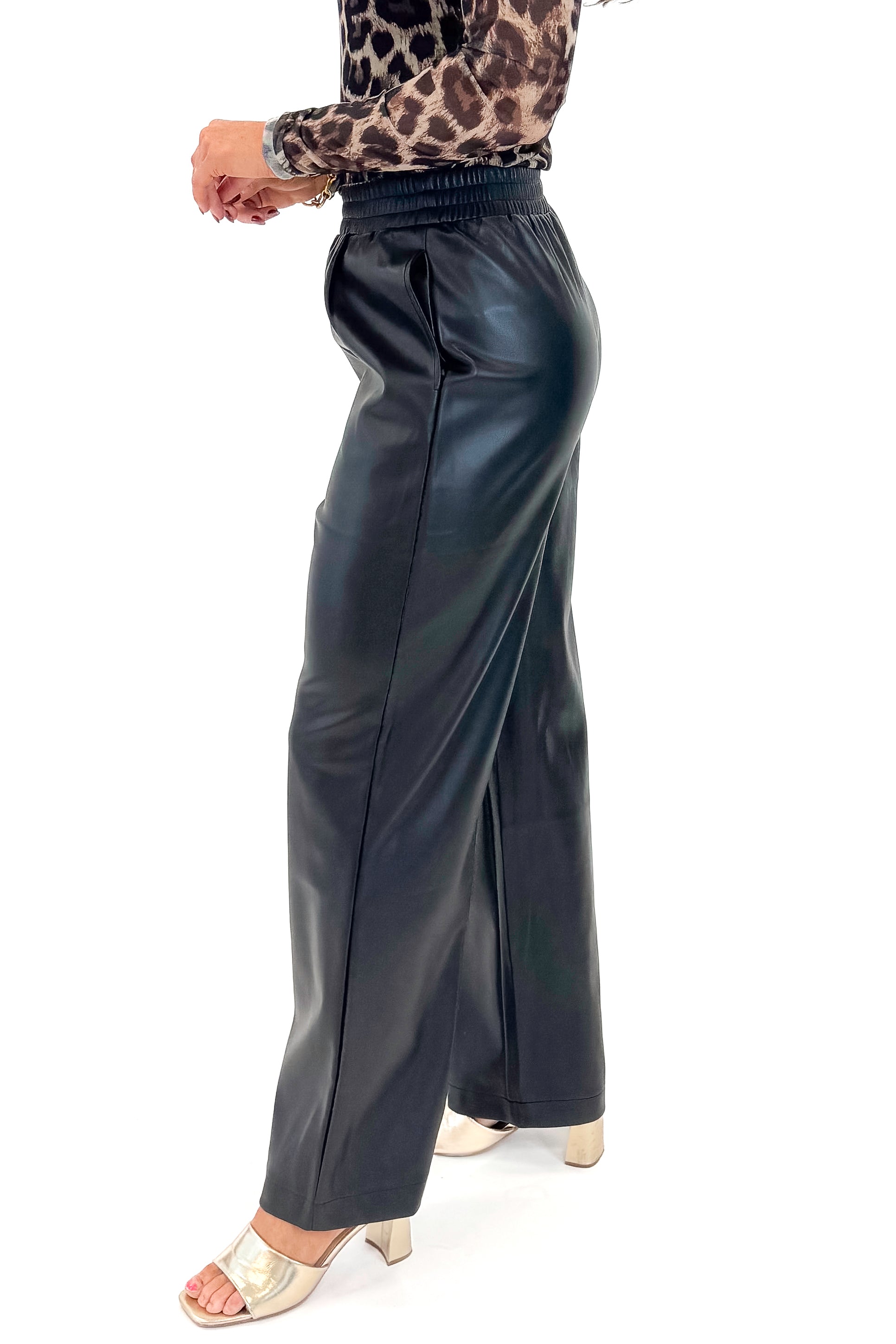 Side detail view of woman wearing Black Faux Leather Pants with leopard mesh top against white studio background.