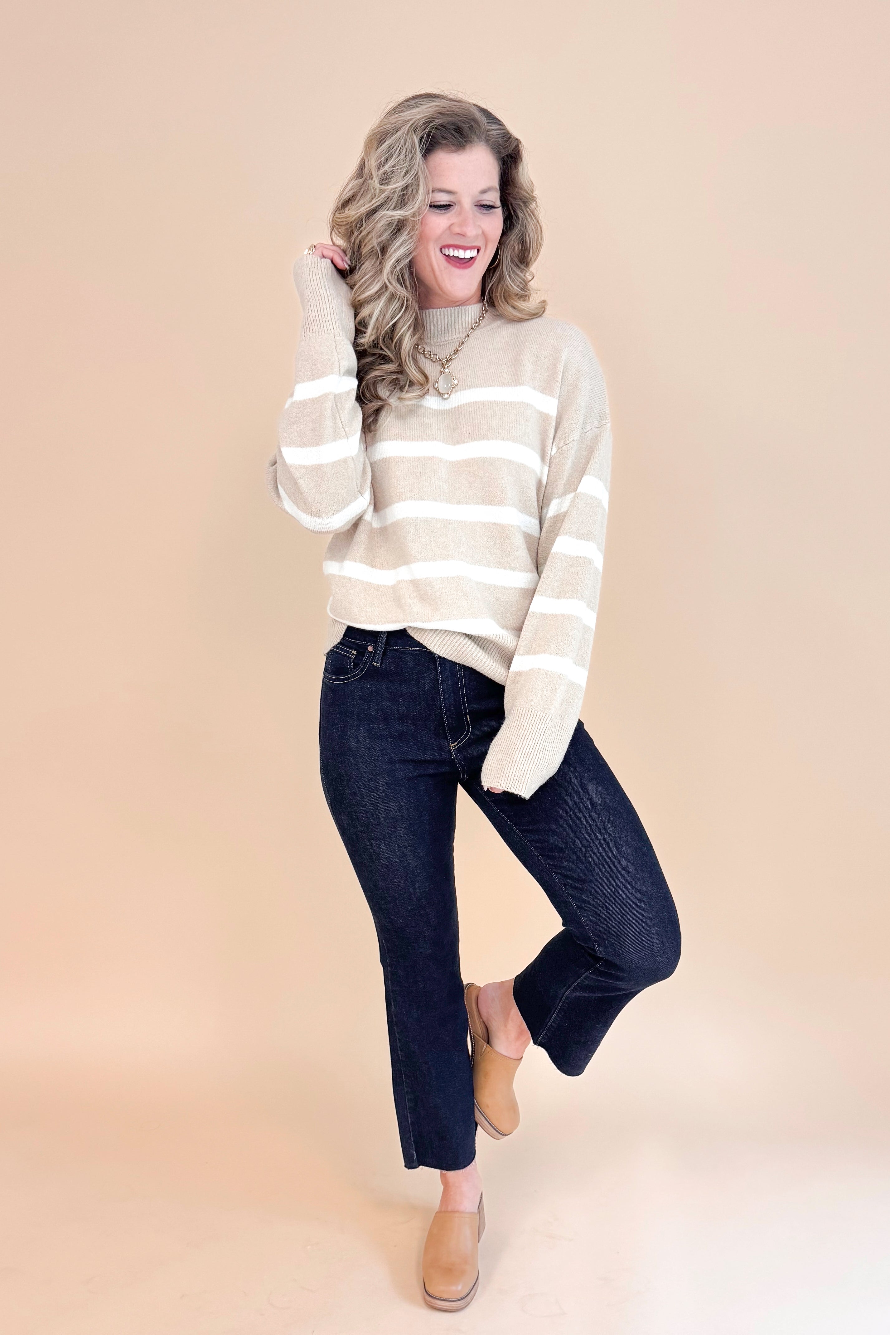 Studio picture of woman wearing Taupe and Cream Striped Mock Neck Sweater and jeans with tan background.