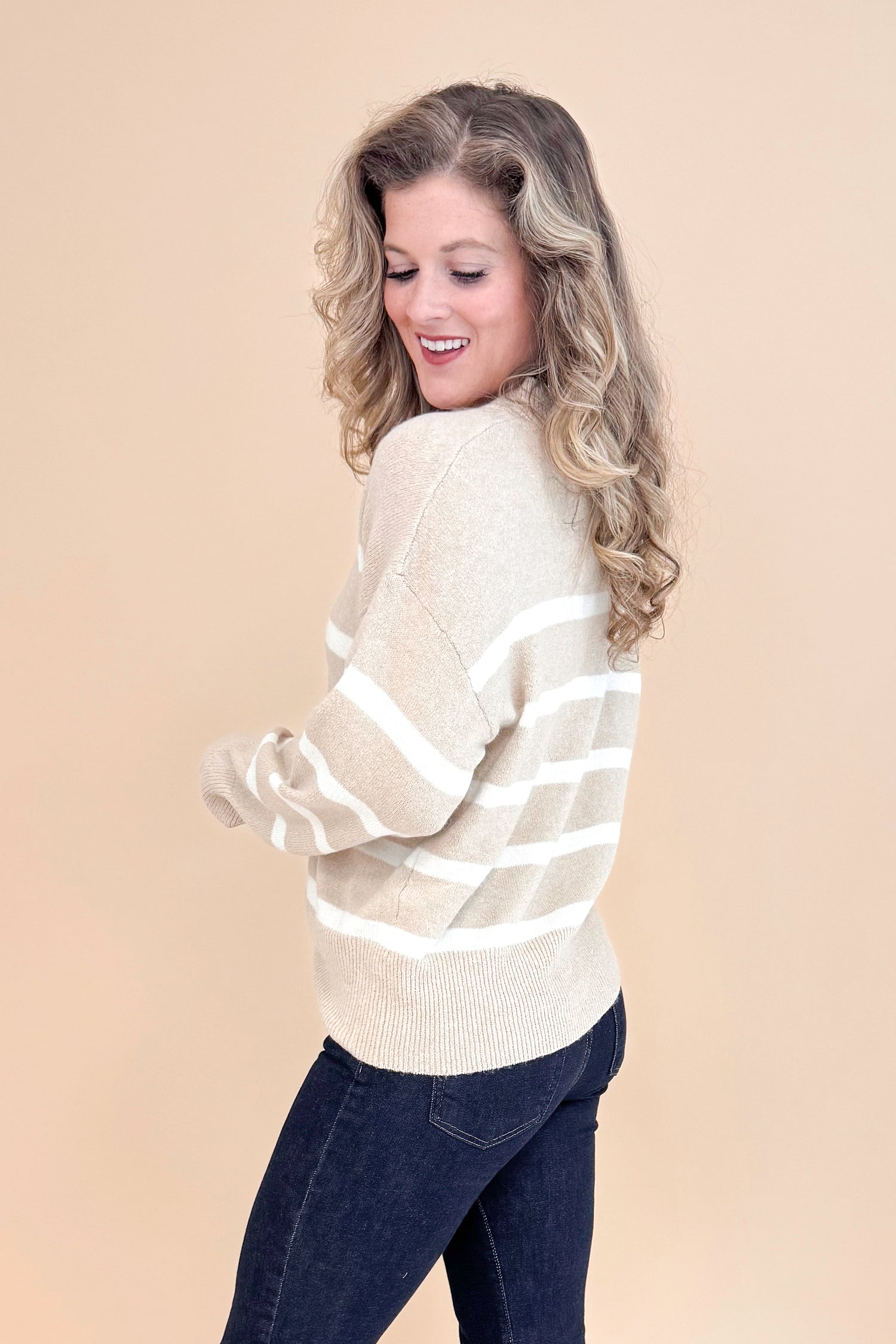 Side view of studio picture of woman wearing Taupe and Cream Striped Mock Neck Sweater and jeans with tan background.