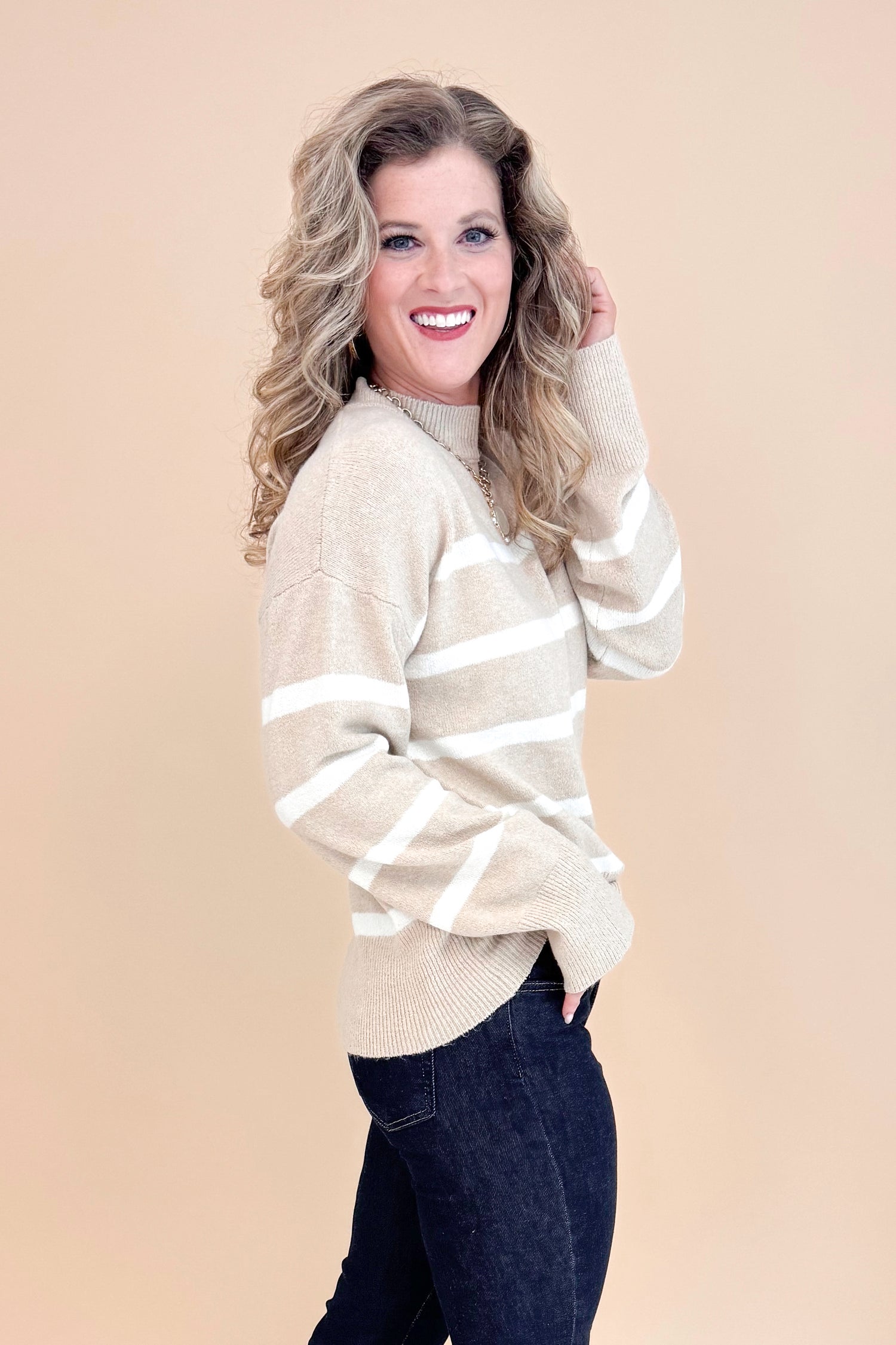 Side view of studio picture of woman wearing Taupe and Cream Striped Mock Neck Sweater and jeans with tan background.