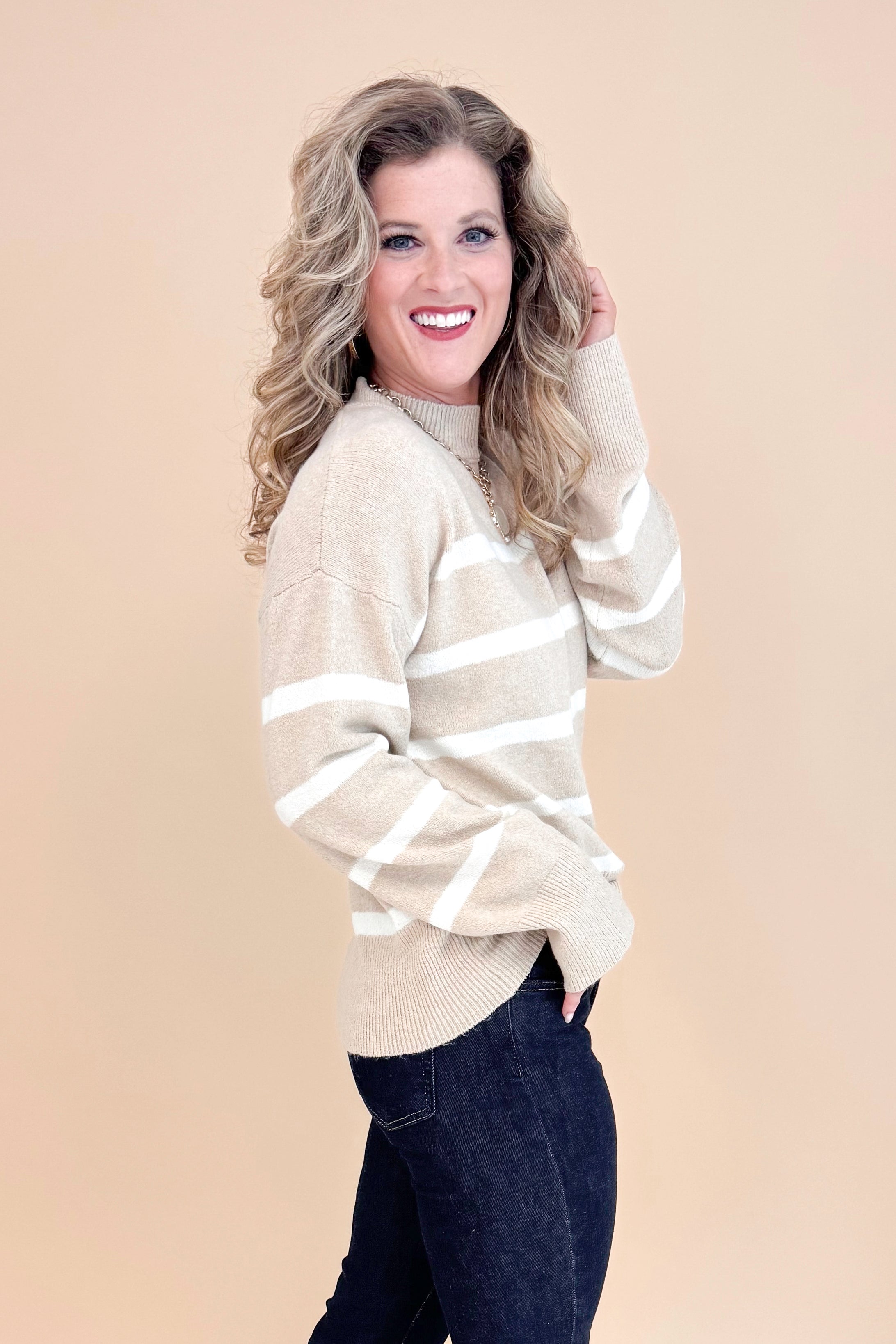 Side view of studio picture of woman wearing Taupe and Cream Striped Mock Neck Sweater and jeans with tan background.