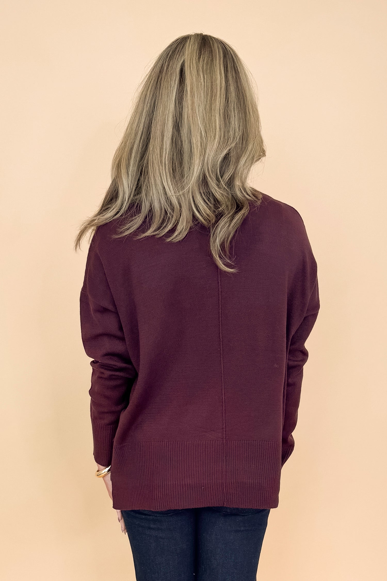 Back view of woman in a studio wearing Wine Center Seam Round Neck Sweater and jeans with a tan background.