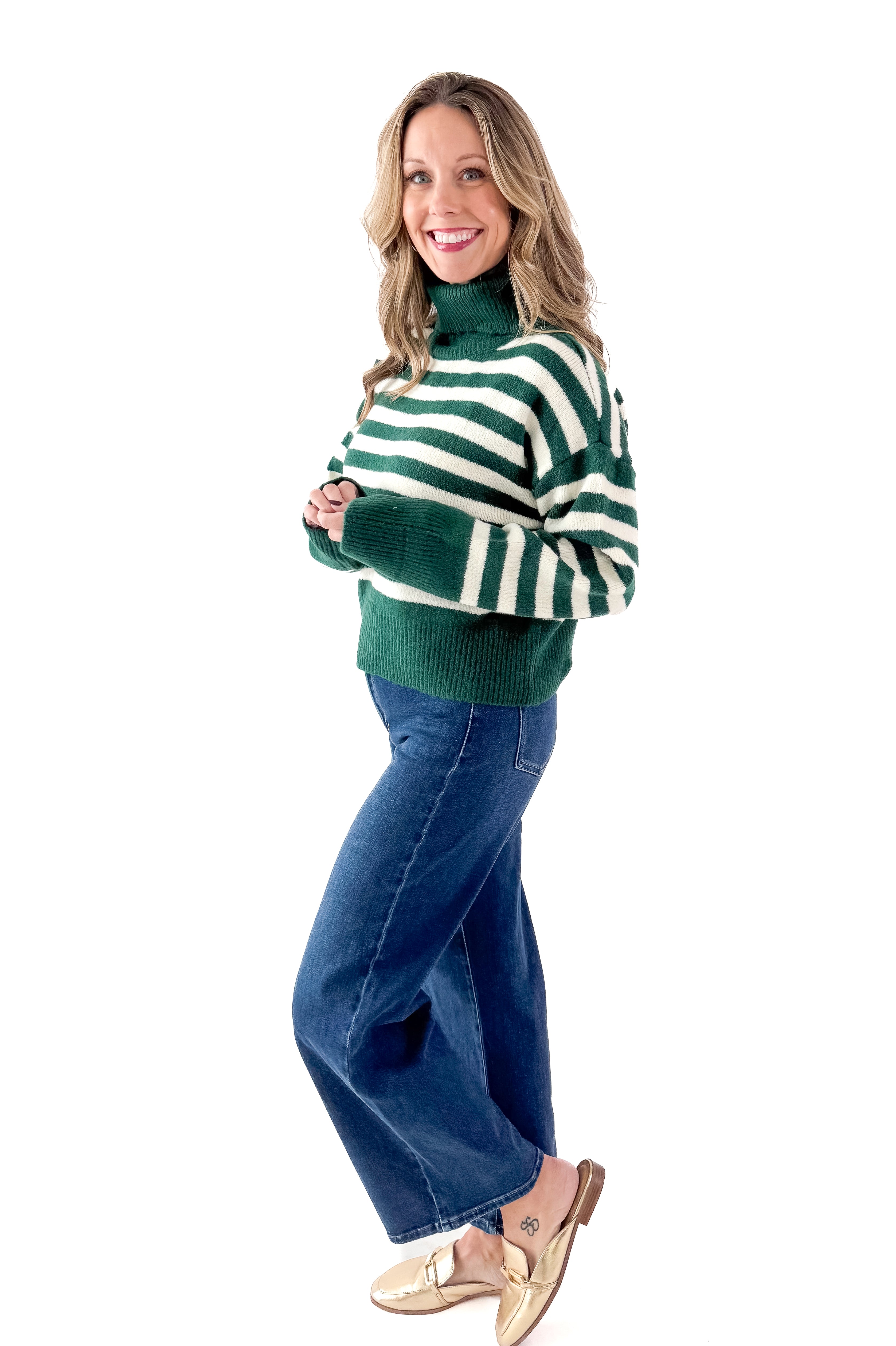 Side view of woman wearing Green & Cream Striped Turtleneck Sweater with jeans against white studio background.