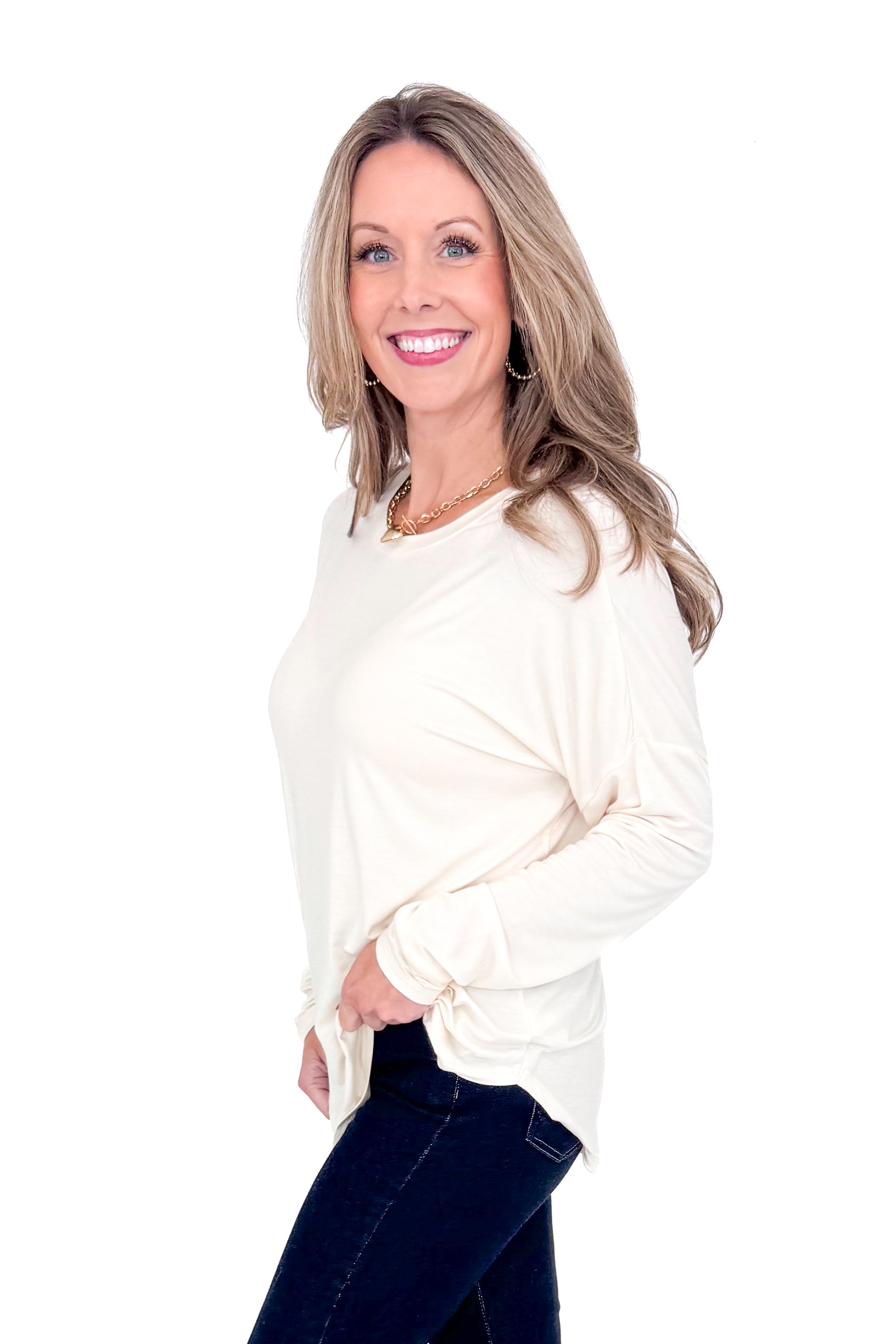 Side view of woman with cream long sleeve curve hem top and blue jeans against white studio background.