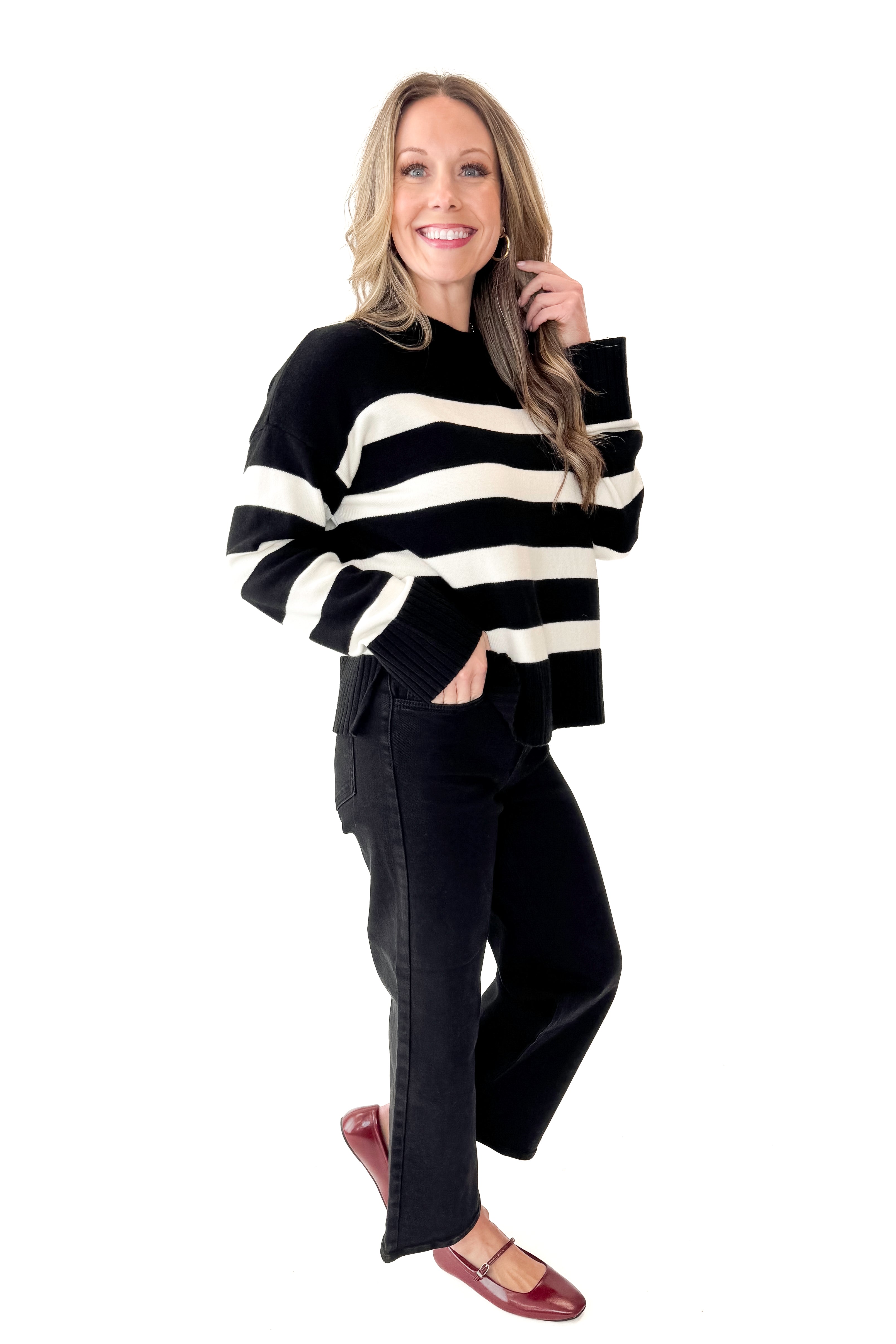 Front view of woman wearing Black and Cream Striped Crewneck Sweater with jeans against white studio background.