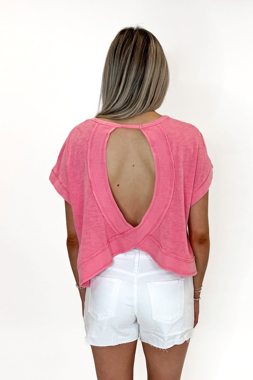 Back view of woman wearing Pink Cut Out Back Tee with jean shorts against white studio background.