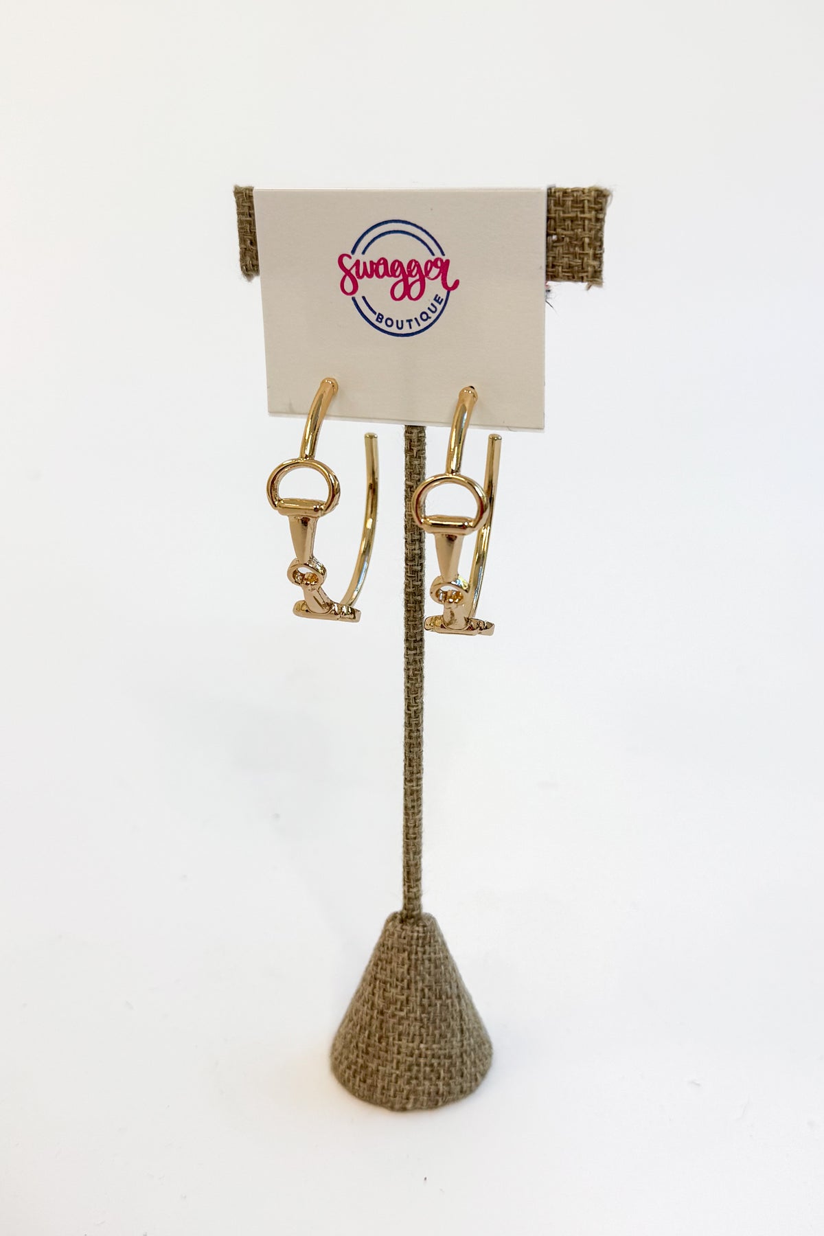 A pair of gold horse-bit style hoop earrings displayed on a textured stand with a branded card that reads &