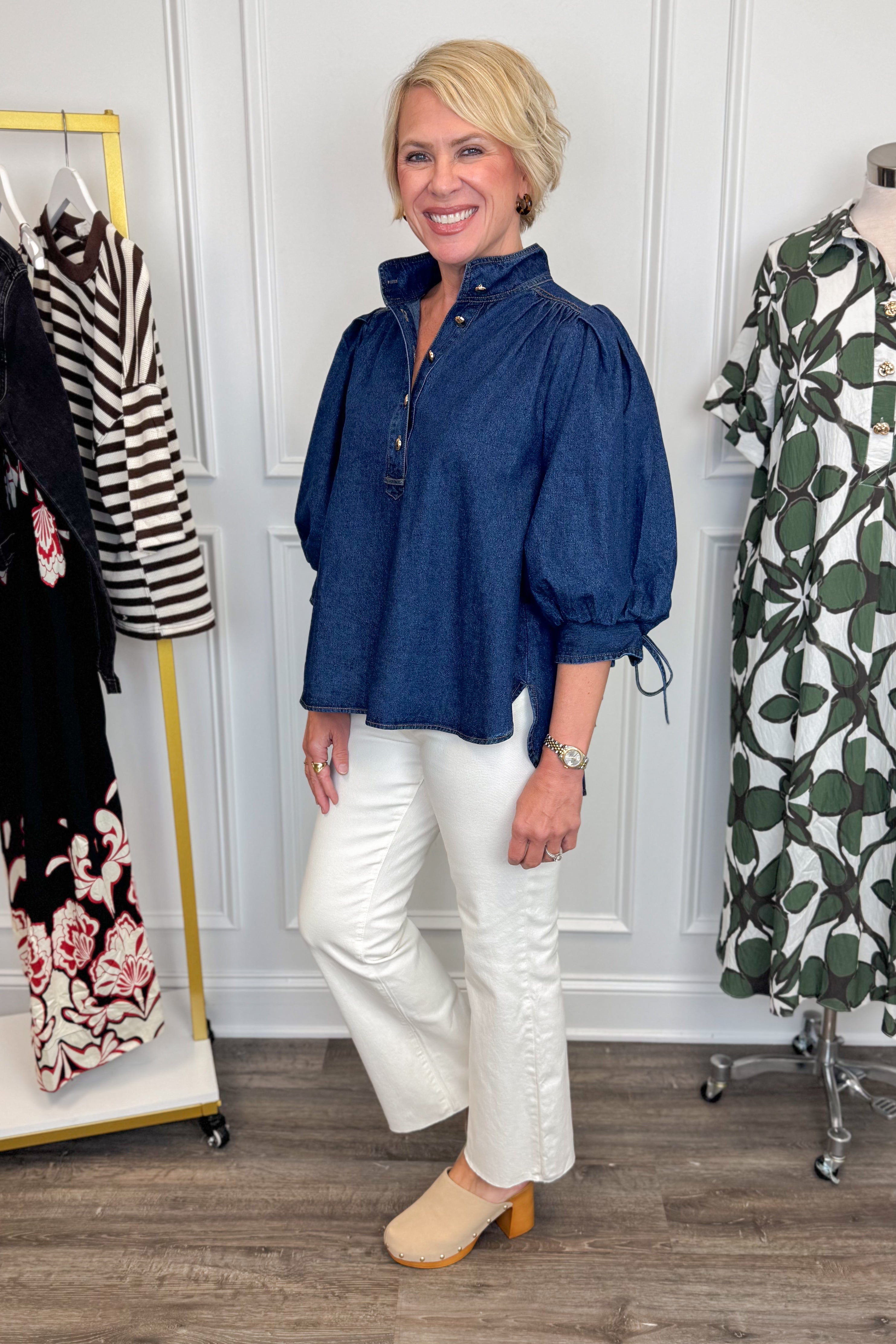 Side view of woman wearing Swagger Boutiqueās Dark Wash Puff Sleeve Denim Top by Entro with jeans with clothing hanging in the background.