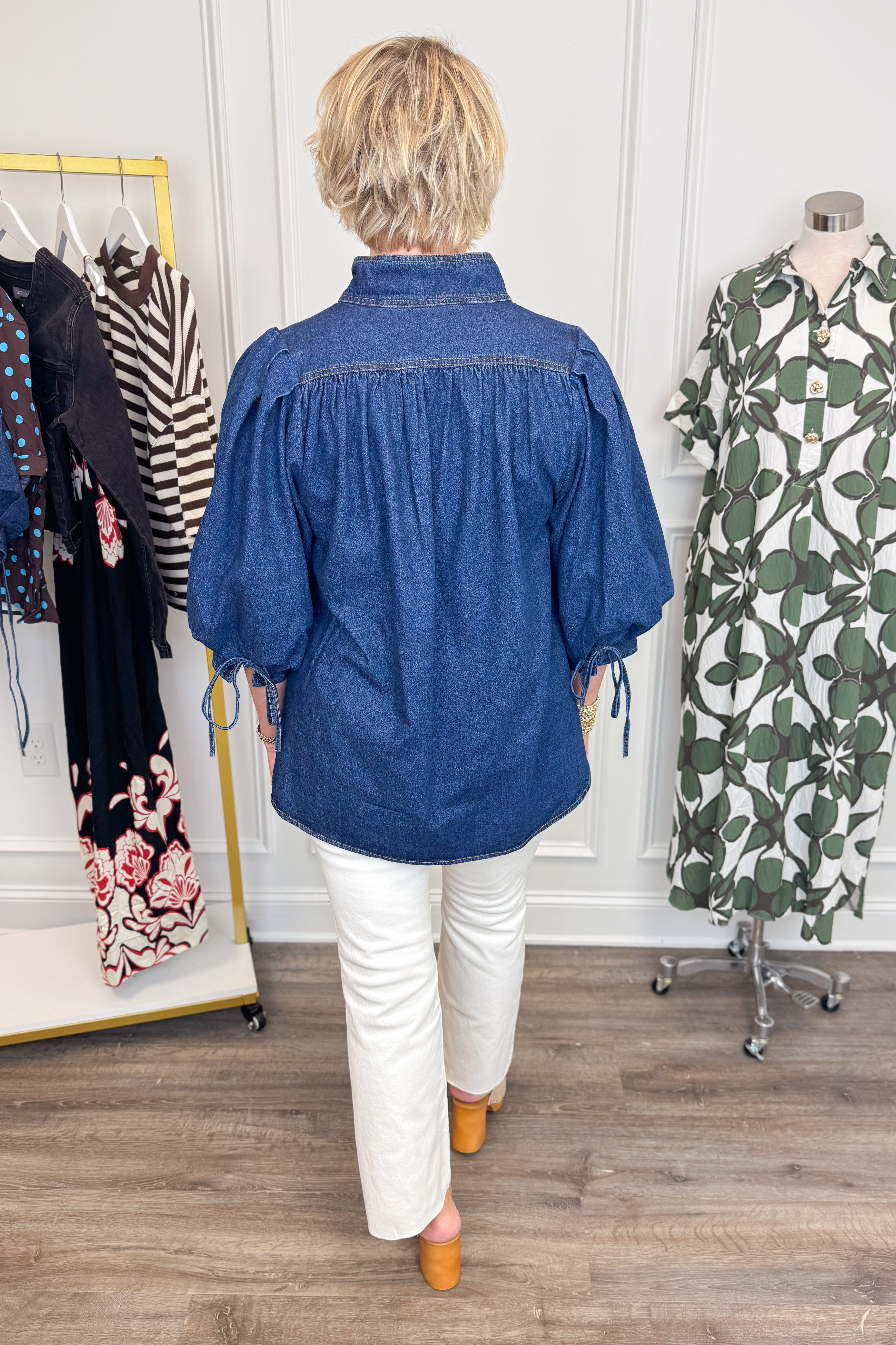 Back view of woman wearing Swagger Boutiqueās Dark Wash Puff Sleeve Denim Top by Entro with jeans with clothing hanging in the background.