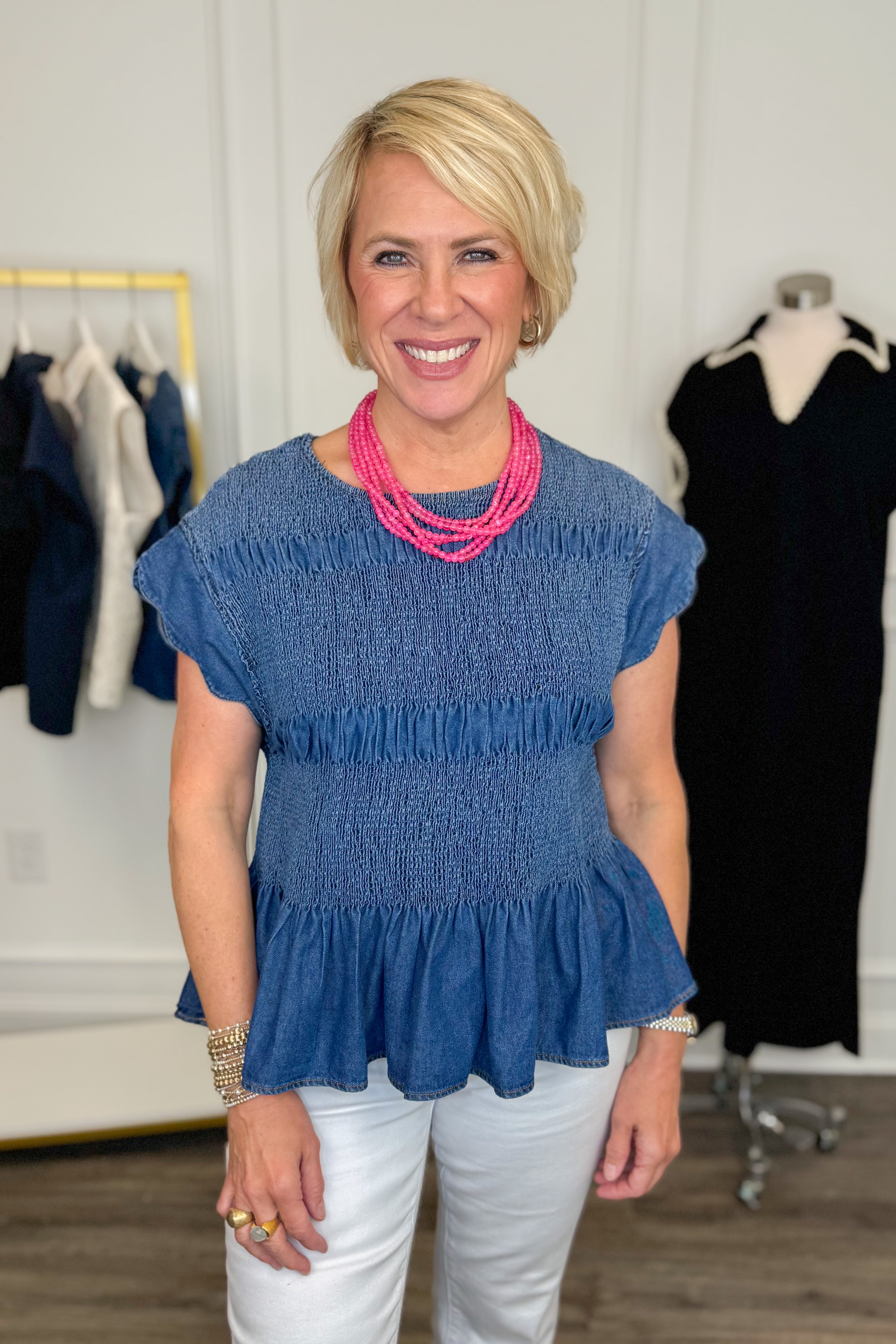 Front view of woman wearing Swagger Boutiqueβs Denim Smocked & Scalloped Top with jeans with clothing hanging in the background.