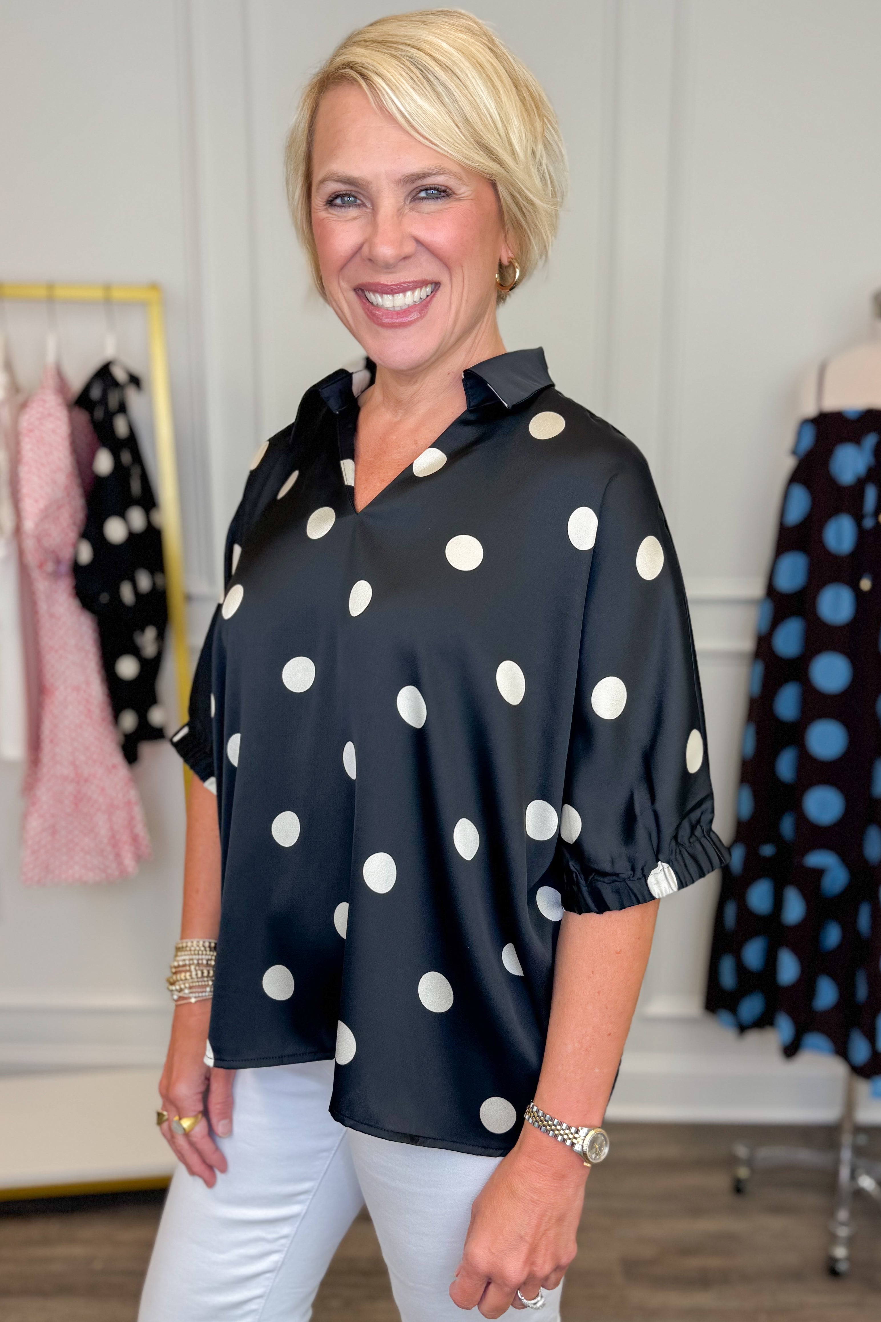Side view of woman wearing Swagger Boutiqueās Black & Cream Polka Dot Satin Top with jeans with clothing hanging in the background.
